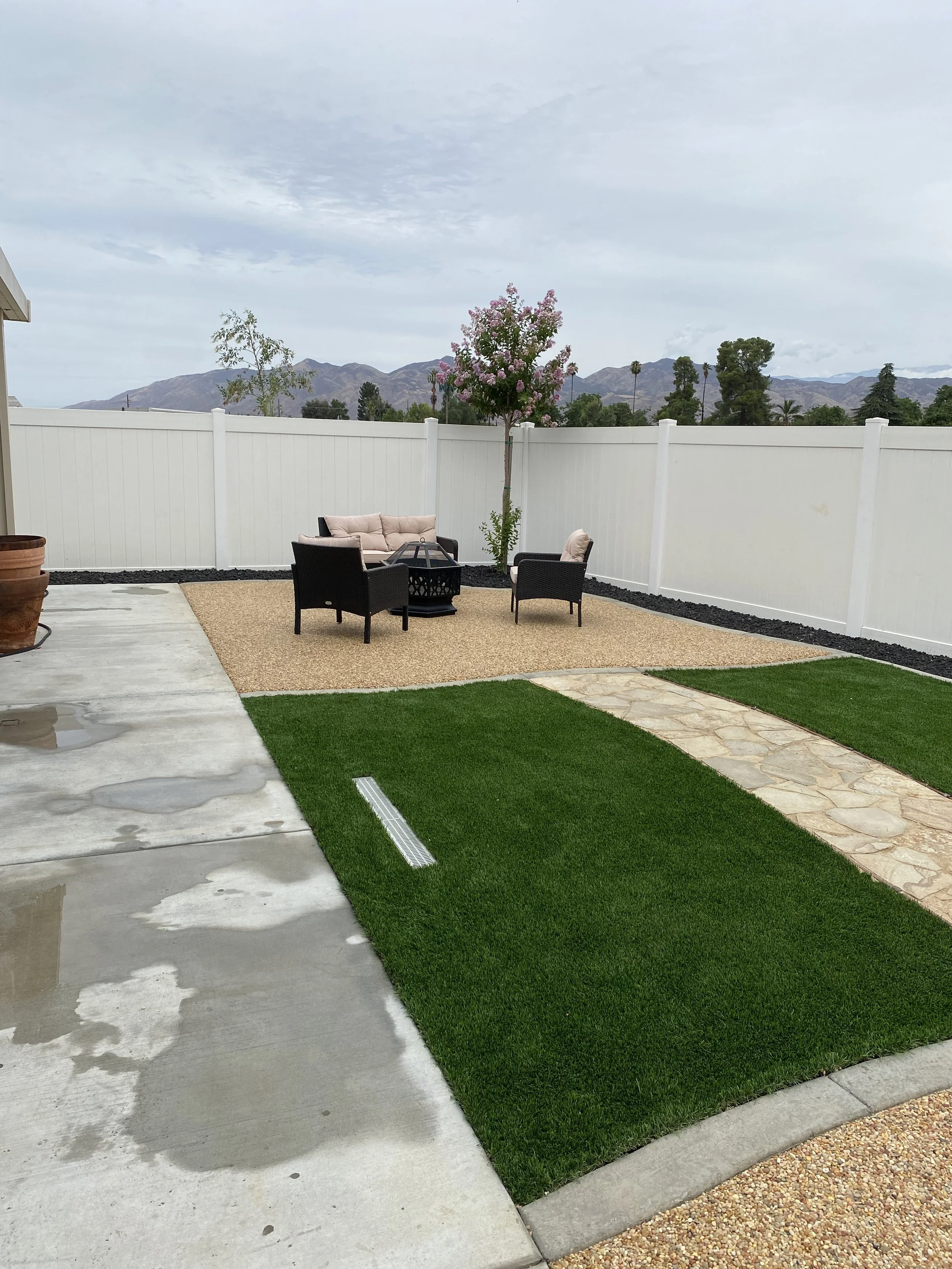 A backyard patio with a white privacy fence, a small tree with pink blossoms, a sitting area with black wicker furniture and cushions, a stepping stone pathway, artificial turf grass, and a mountain landscape in the background.