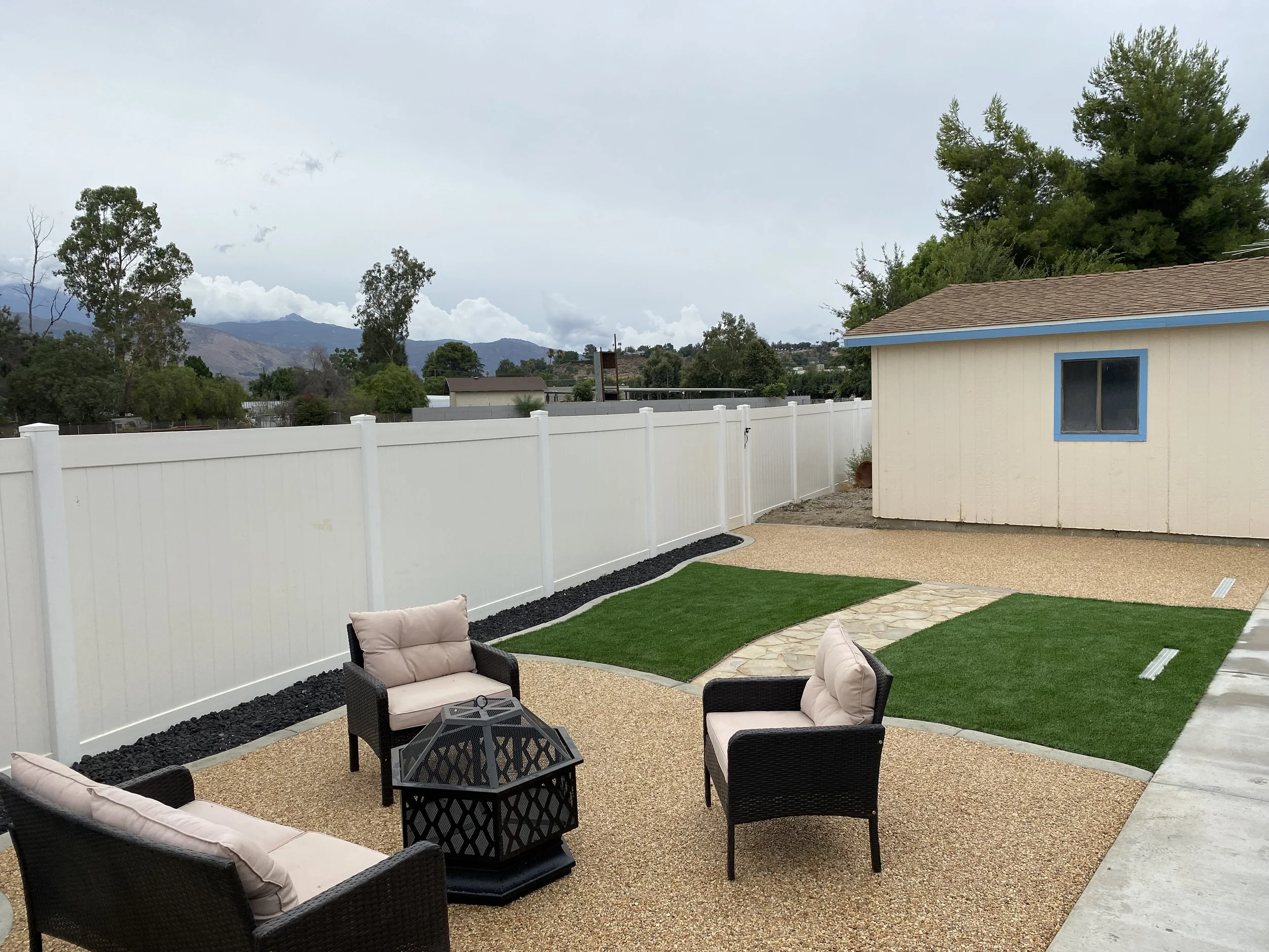 A backyard patio with beige cushioned outdoor chairs around a fire pit, on a gravel surface, with a small patch of artificial grass, a stone pathway, and a white fence with trees and mountains in the background.