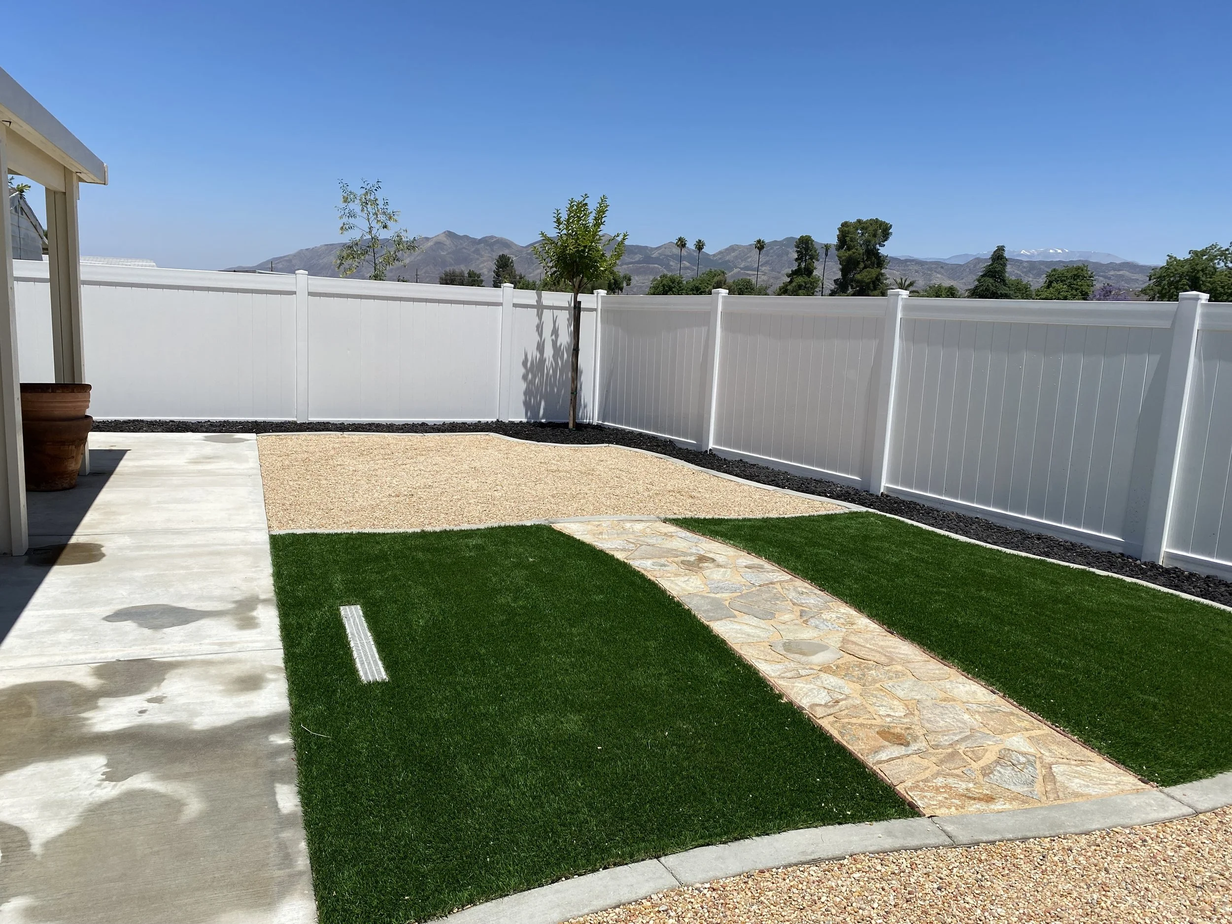 Backyard with a concrete patio, artificial grass, stone pathway, and white privacy fence, with mountain view in the background under clear blue sky.