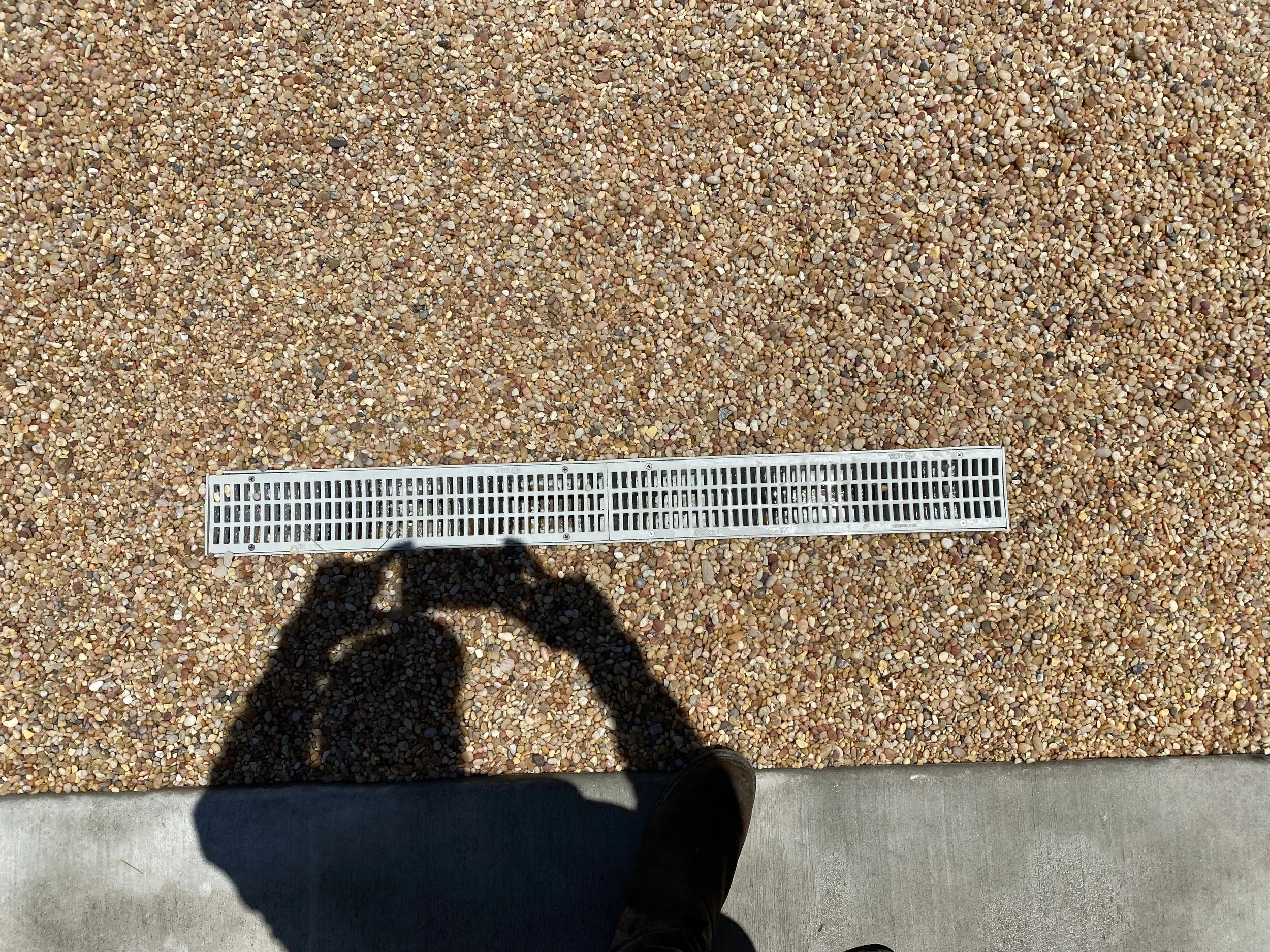 A gravel surface with a metal drain cover and a shadow of a person taking a photo, with a concrete sidewalk at the bottom.