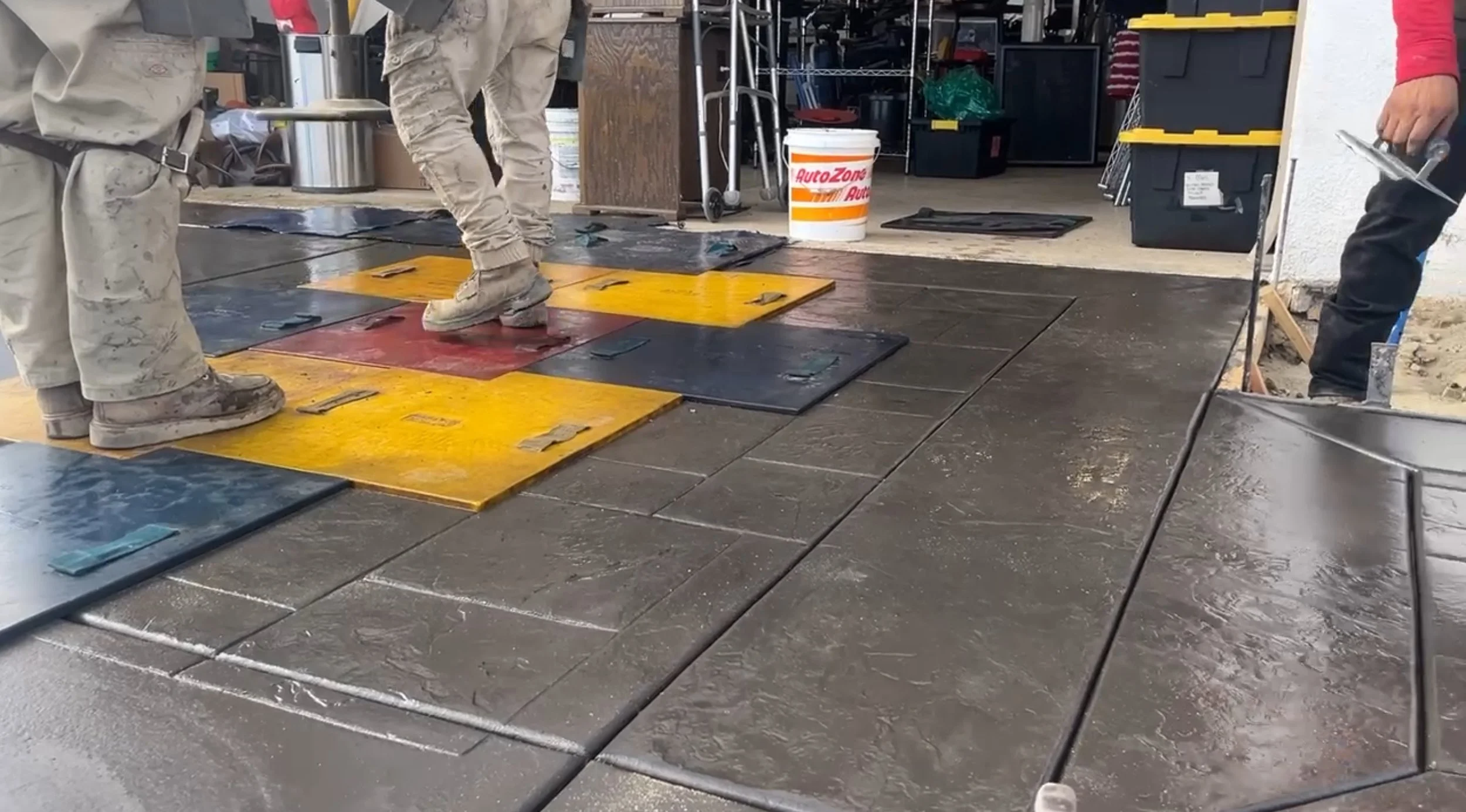 Construction workers installing large interlocking tiles on a concrete floor in a garage or workshop setting. The tiles are in various colors including yellow, red, and dark blue. Construction tools and supplies are visible in the background.