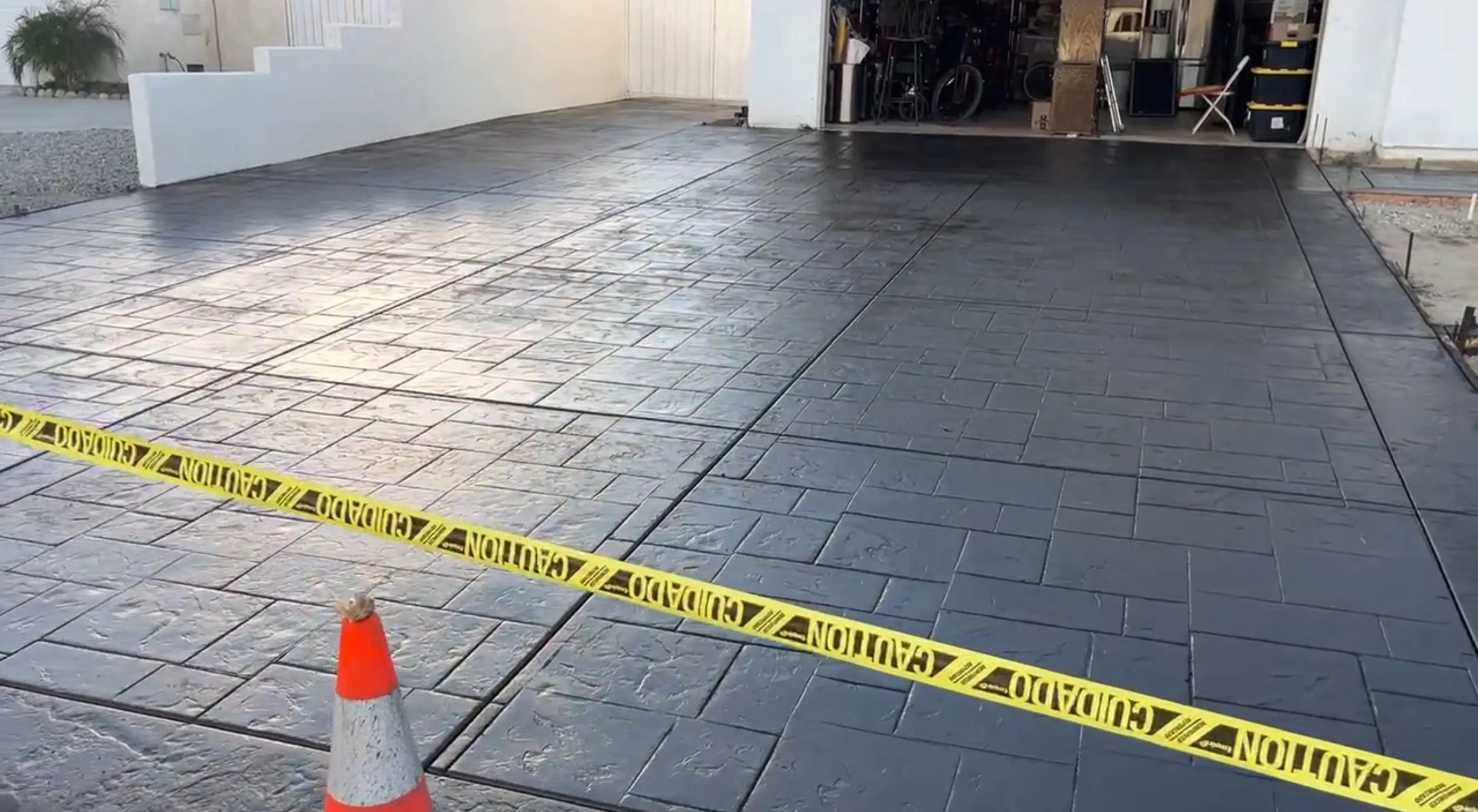 Freshly poured concrete driveway with stamped pattern, bordered by a white wall and a garage filled with tools and equipment, caution tape and an orange traffic cone in the foreground.