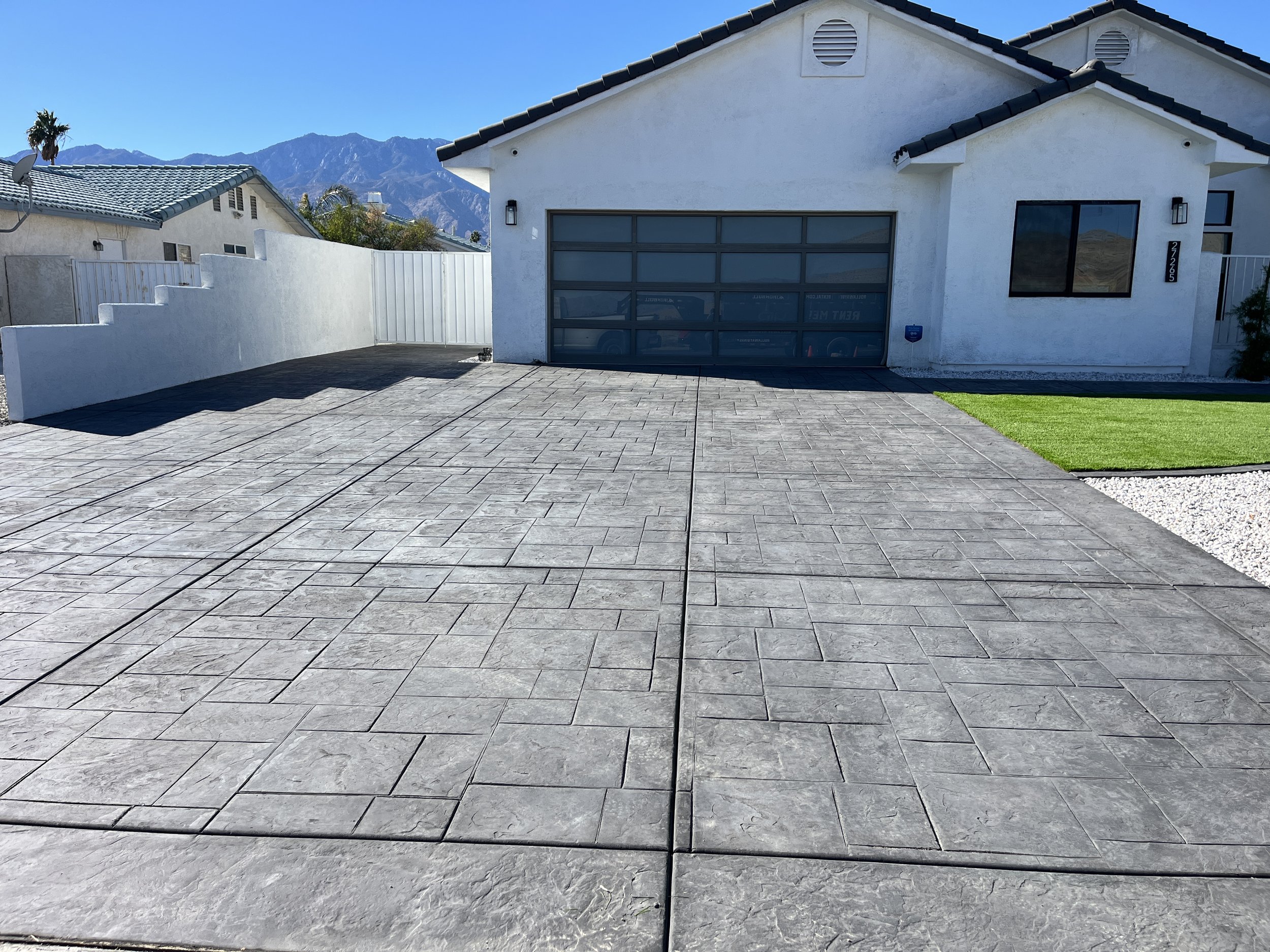 Front view of a white house with a concrete driveway, garage door, and a small patch of green grass on the right, with mountains in the background under a clear blue sky.