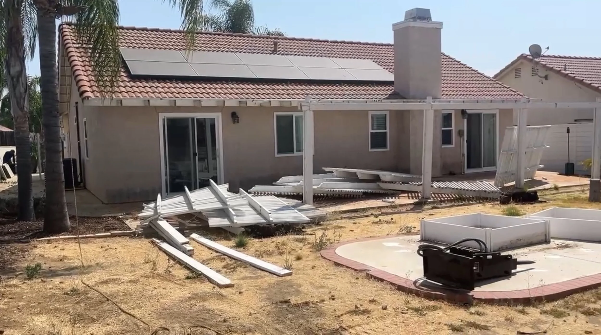 Backyard of a house under renovation with construction materials and damaged structures, including a fallen white fence, a pile of white fence pieces on the ground, and an empty garden bed, with neighboring house and palm trees visible.