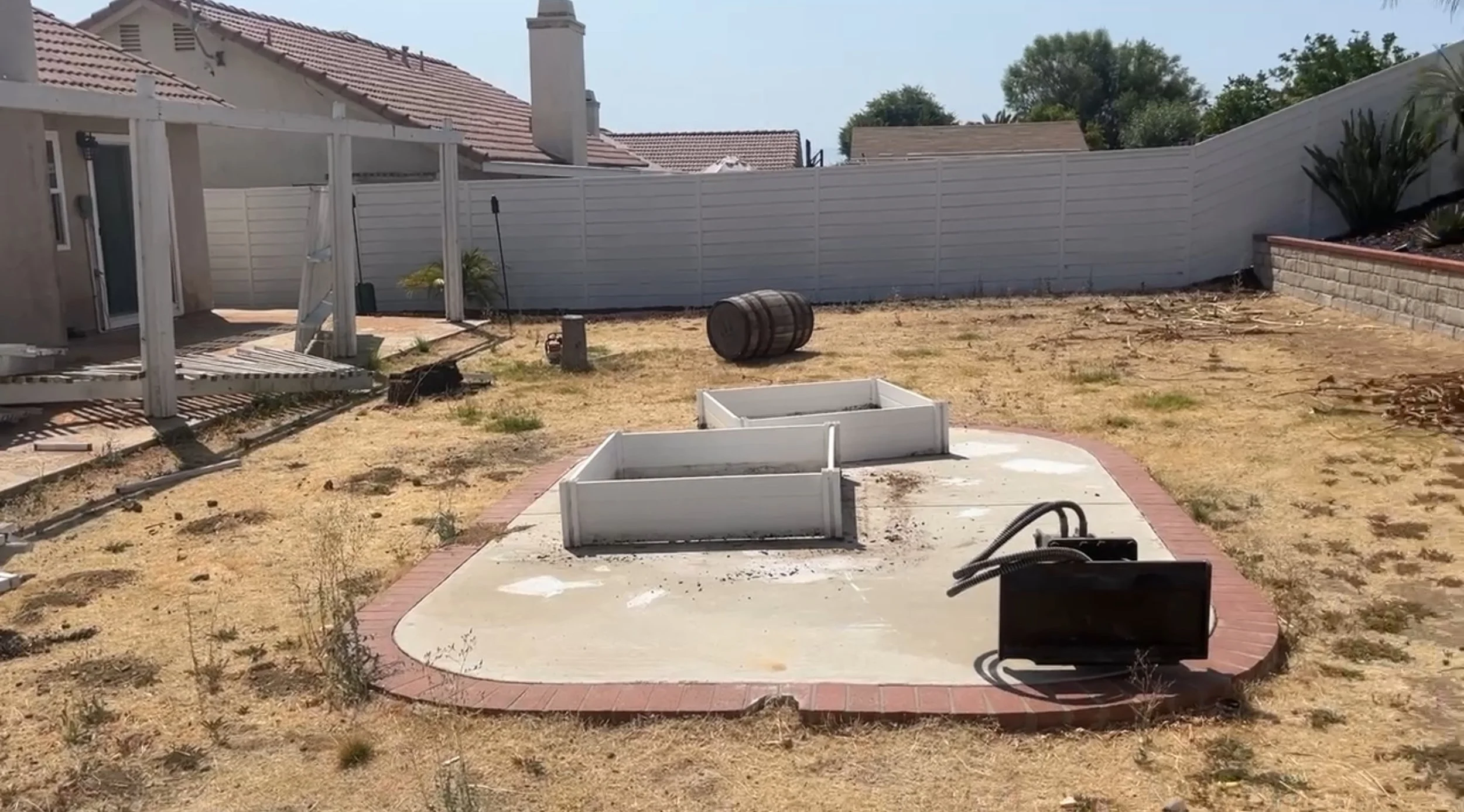 A backyard under construction with a concrete patio, white raised garden beds, a barrel, and a black electrical box. The yard is dry with patches of grass, enclosed by a white fence, with neighboring houses visible.