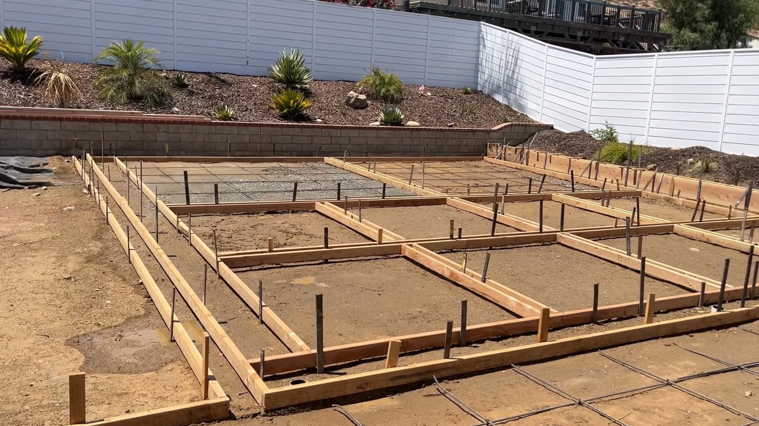 Construction site with wooden framing for concrete slabs, surrounded by a landscaped yard with plants and a white fence.