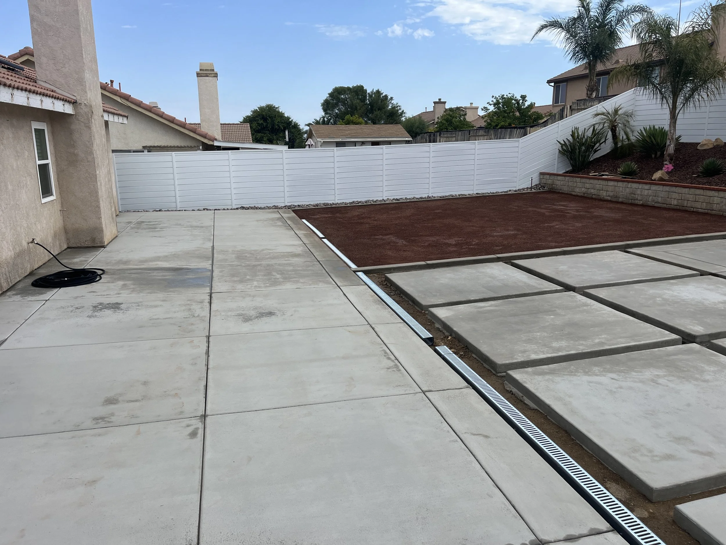 Backyard with concrete patio, garden beds with red soil, and a white privacy fence. There are some plants and small trees near the garden beds, and neighboring houses are visible in the background.