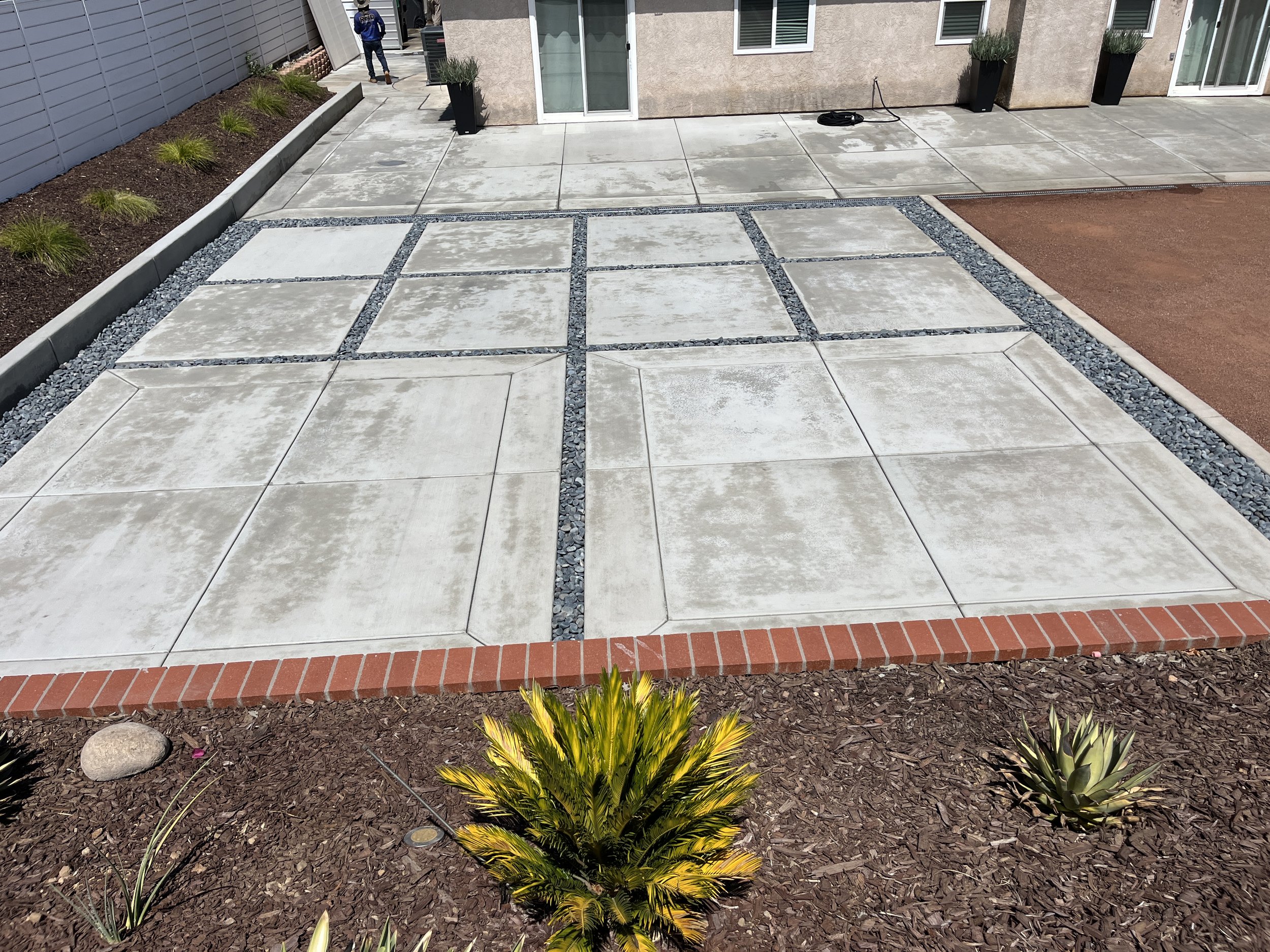 A newly constructed patio with concrete slabs divided by gravel strips in front of a modern house. Potted plants are on the patio and a man is walking in the background.