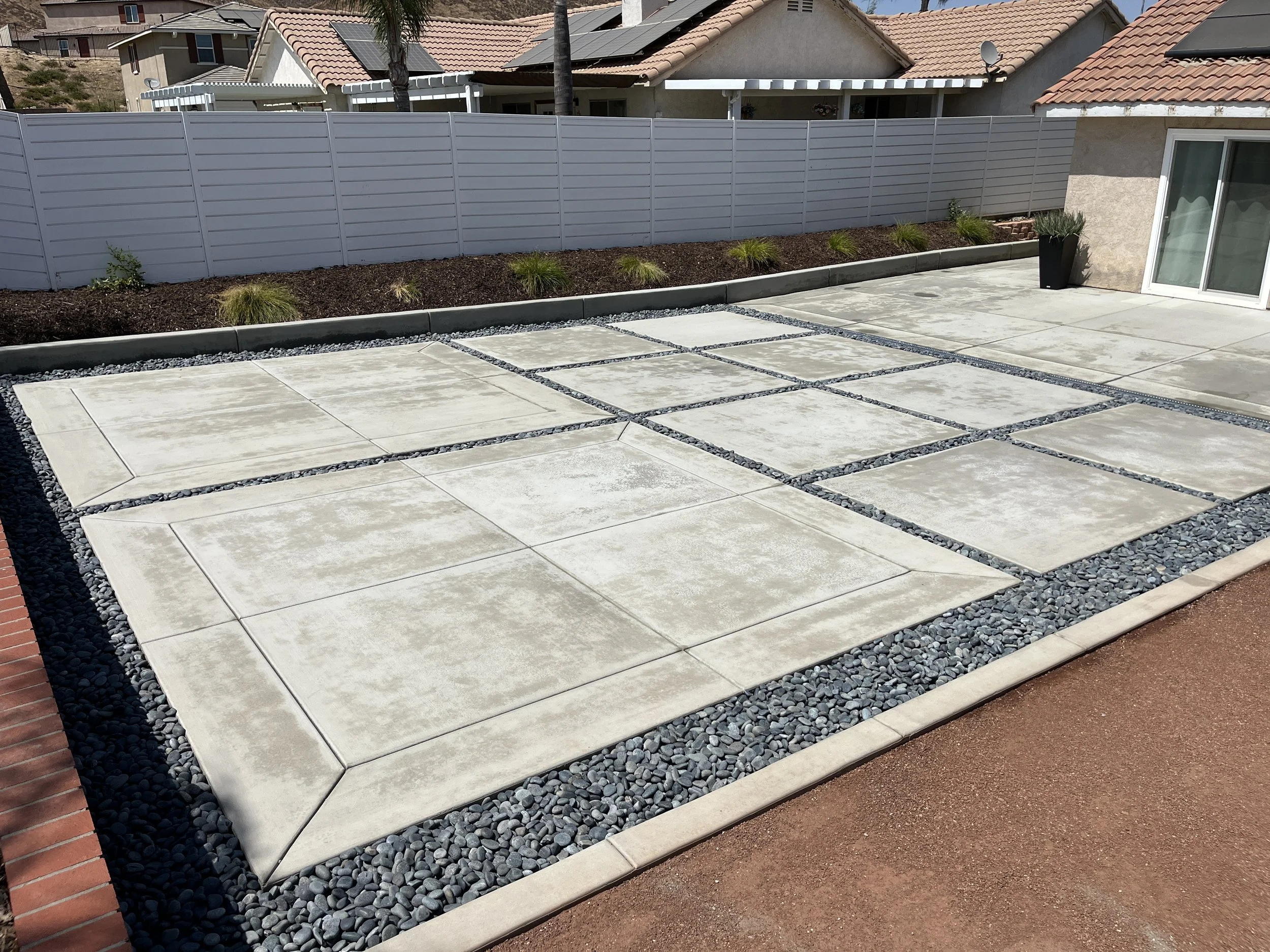 A backyard patio with concrete square slabs separated by small black pebbles, surrounded by a white fence and garden bed with plants, next to a house with sliding glass door and potted plants.