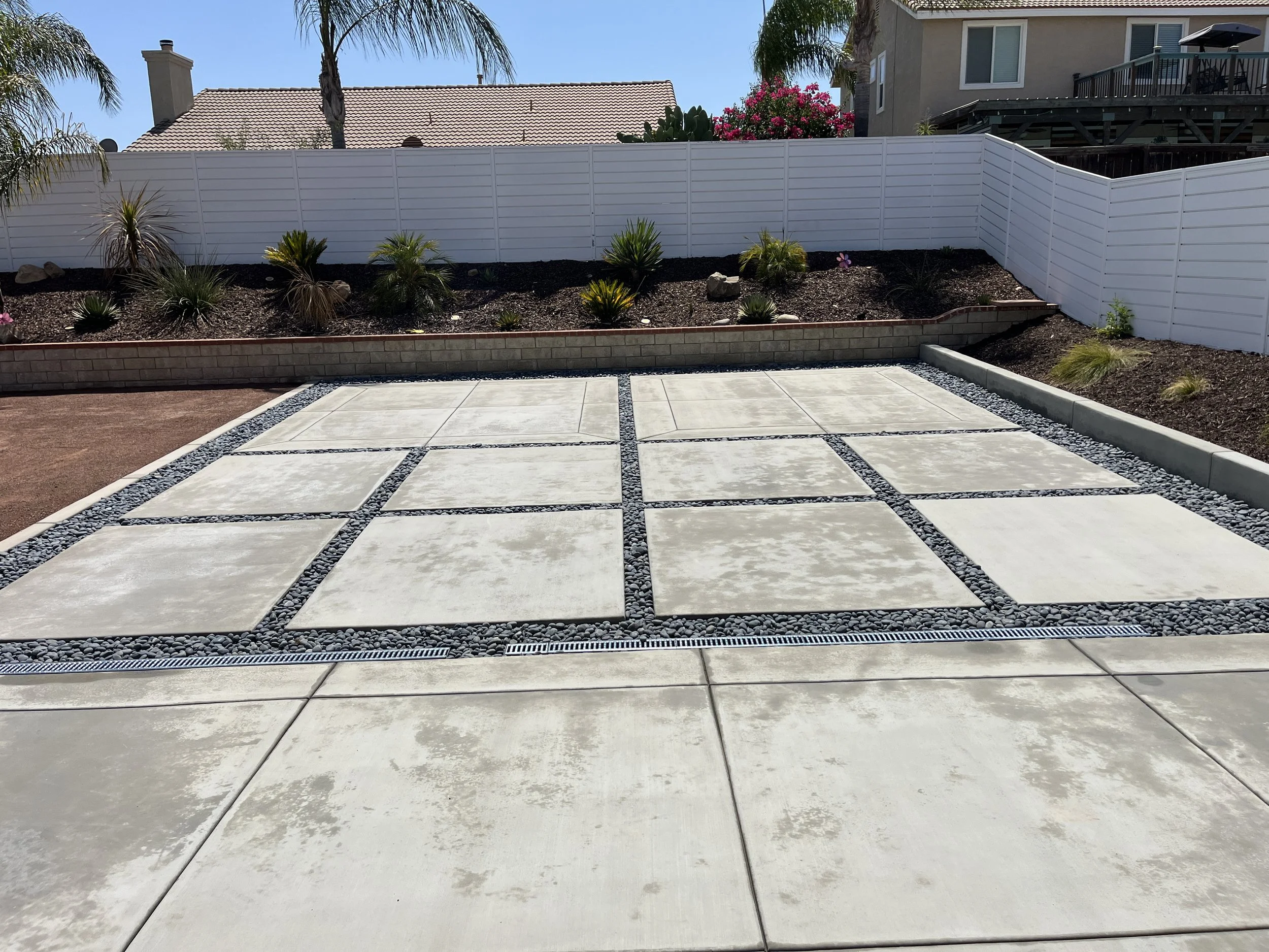 A modern outdoor patio with large concrete tiles separated by small black rocks, bordered by white edging and a gravel strip. There is a landscaped area with shrubs, plants, and a white privacy fence in the background.