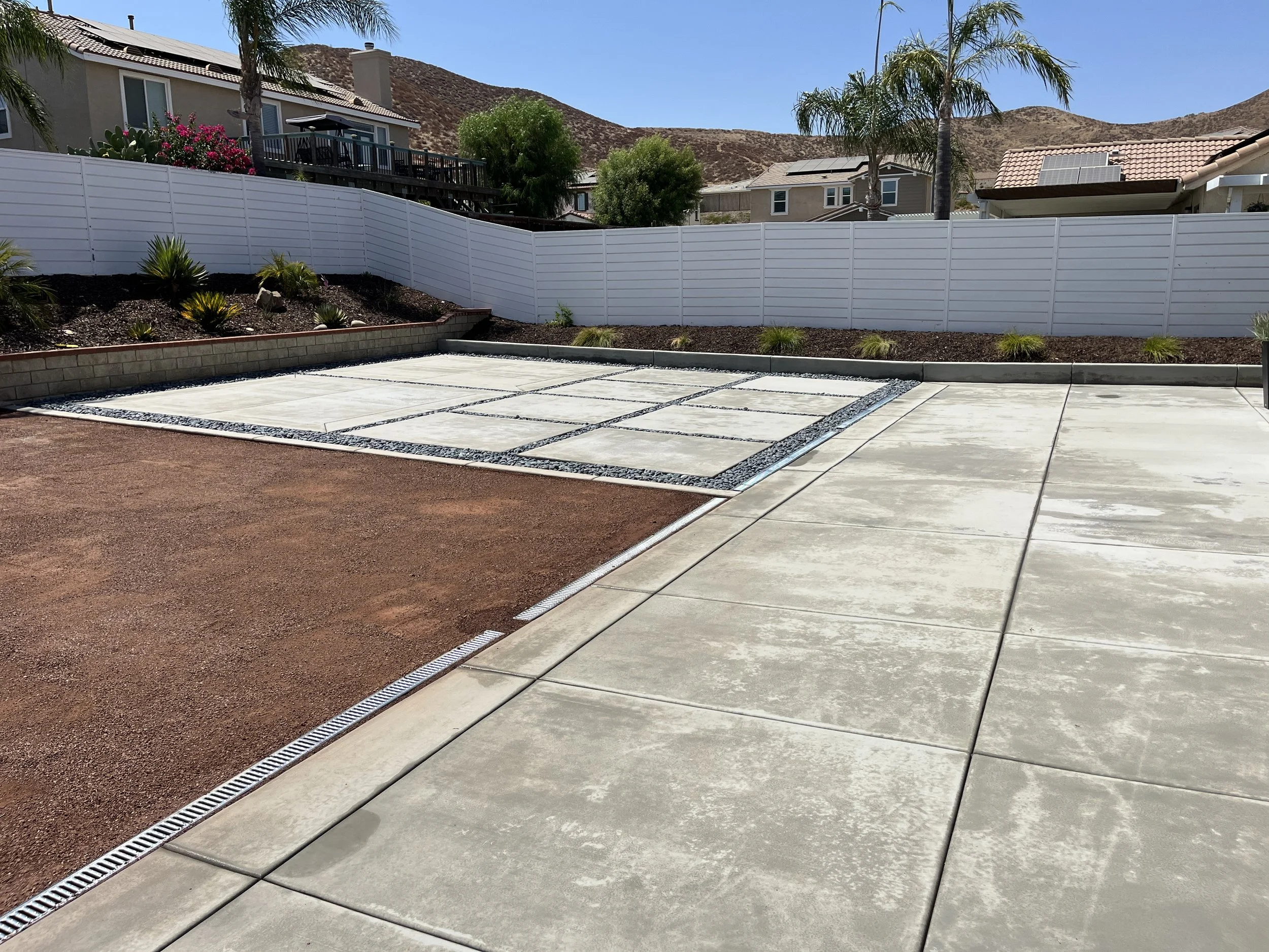 A backyard with a paved concrete walkway, a red dirt area, and a new section with concrete pavers separated by gravel. There are small plants along the back fence, which is white and made of panels. There are houses and hills in the background with palm trees and a clear blue sky.