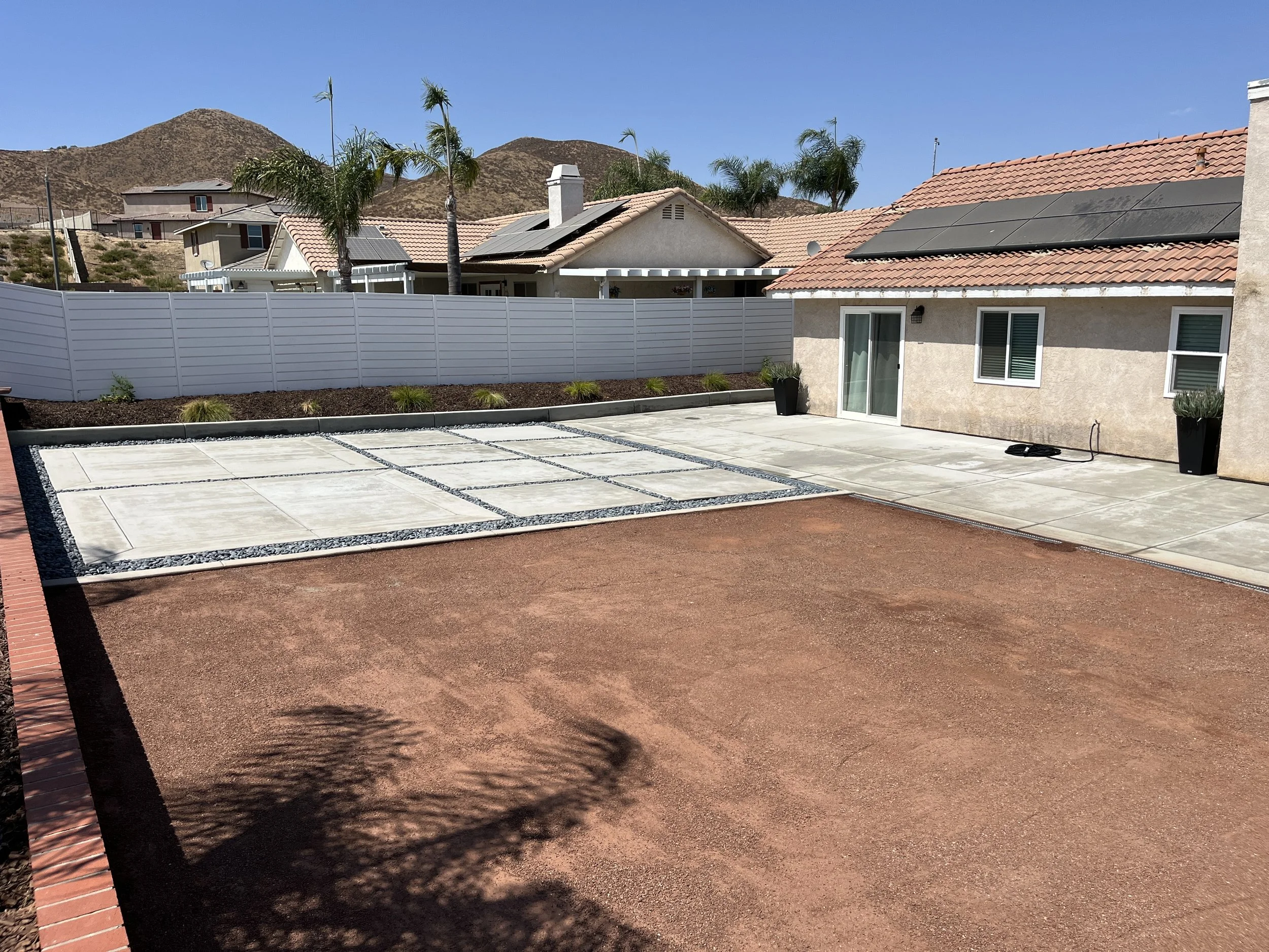 Fully landscaped backyard with a concrete patio, potted plants, and decorative concrete and gravel areas, surrounded by a white privacy fence, with neighboring houses and hills in the background.