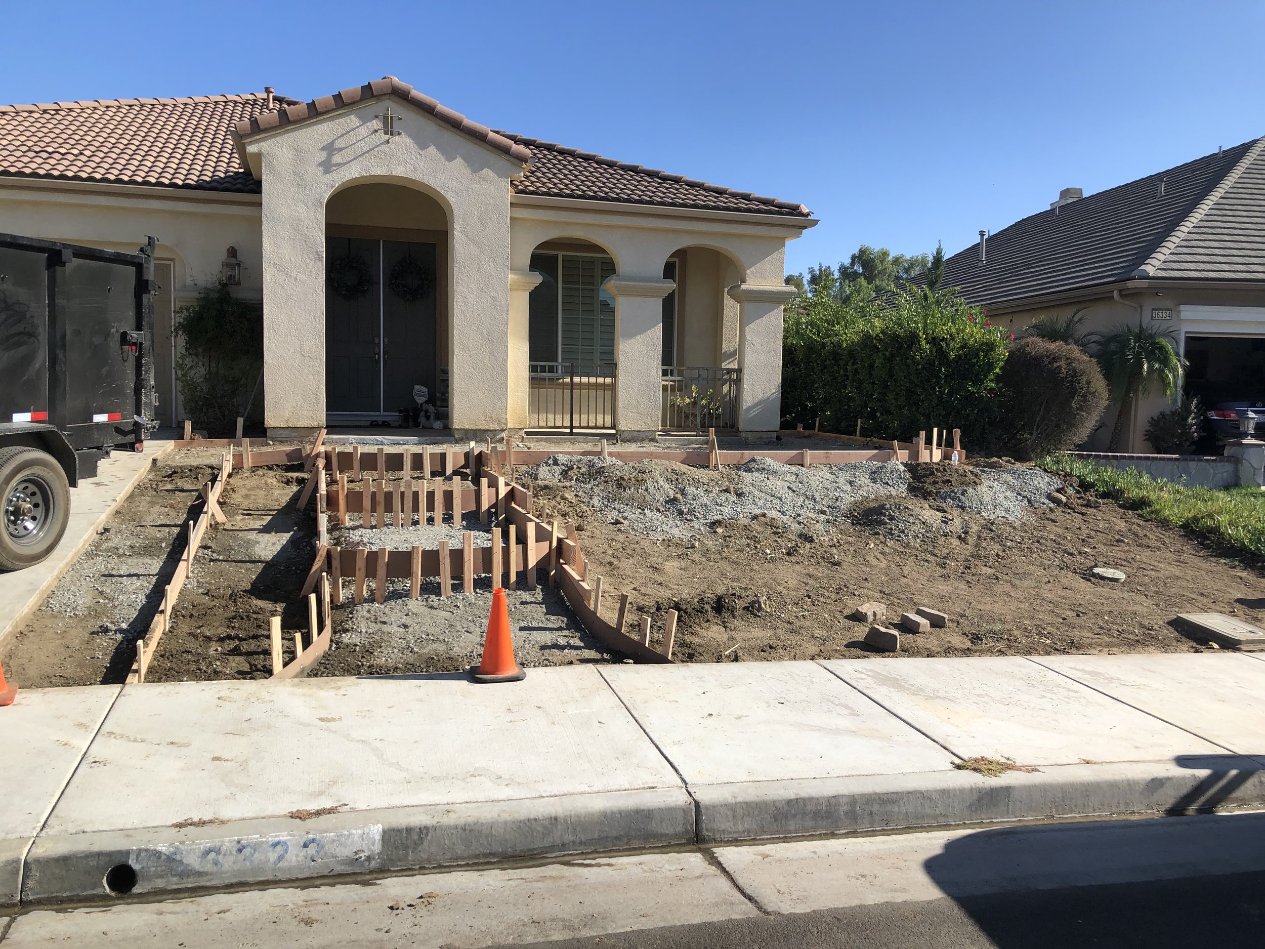 Front yard of a house under construction, with wooden frames for a walkway or garden beds, construction cones, and dirt patches.