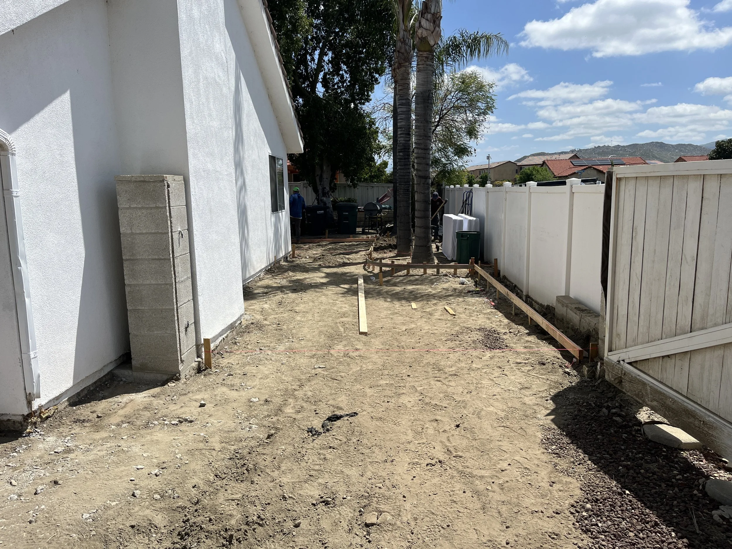 A backyard area with dirt and construction framing, showing a house on the left, a white fence on the right, trees, trash bins, and construction materials under a partly cloudy sky.