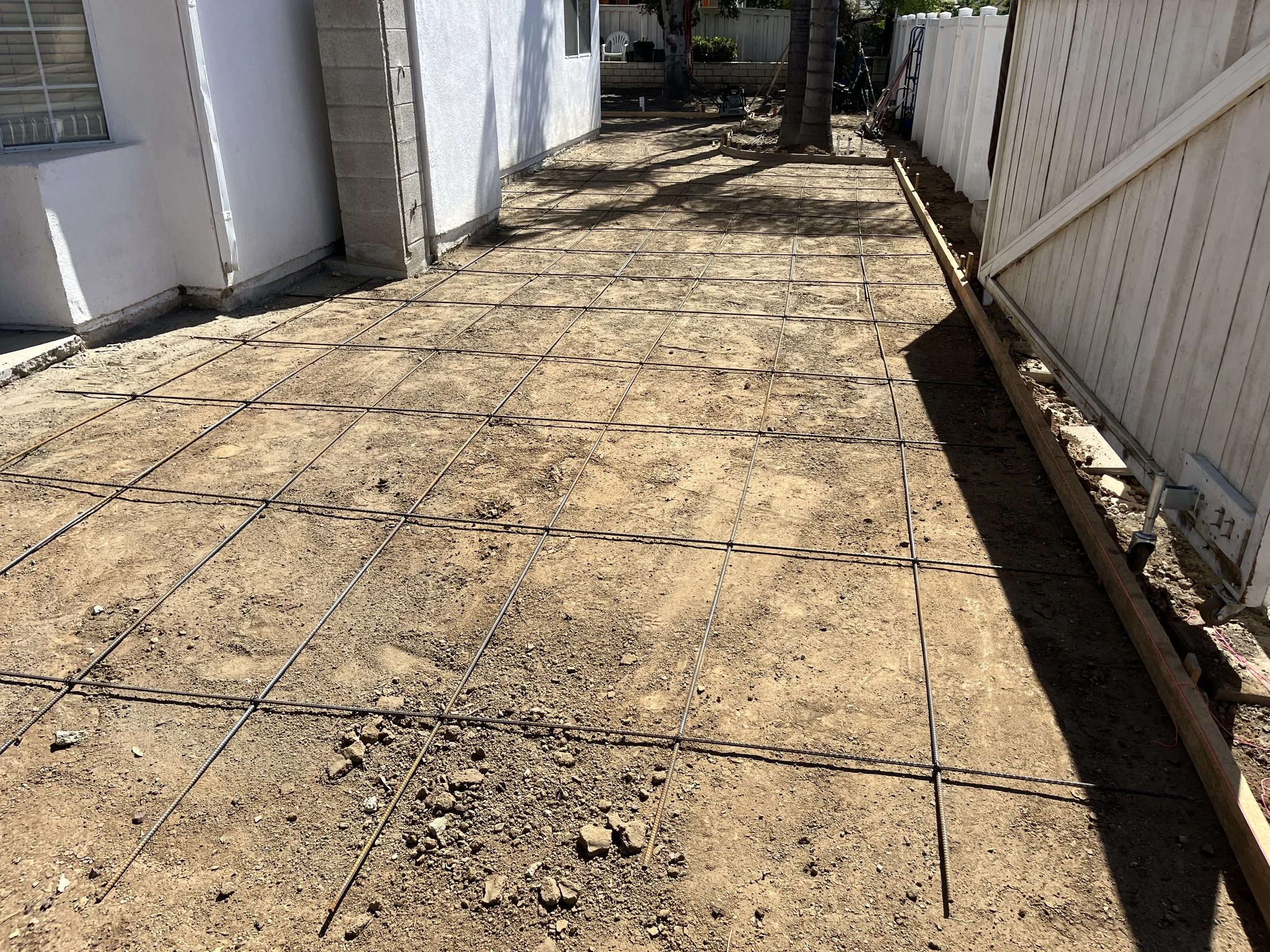 A backyard with a simple fence, showing a newly prepared dirt ground with a grid of metal rebar for concrete pouring, and a few trees in the background.