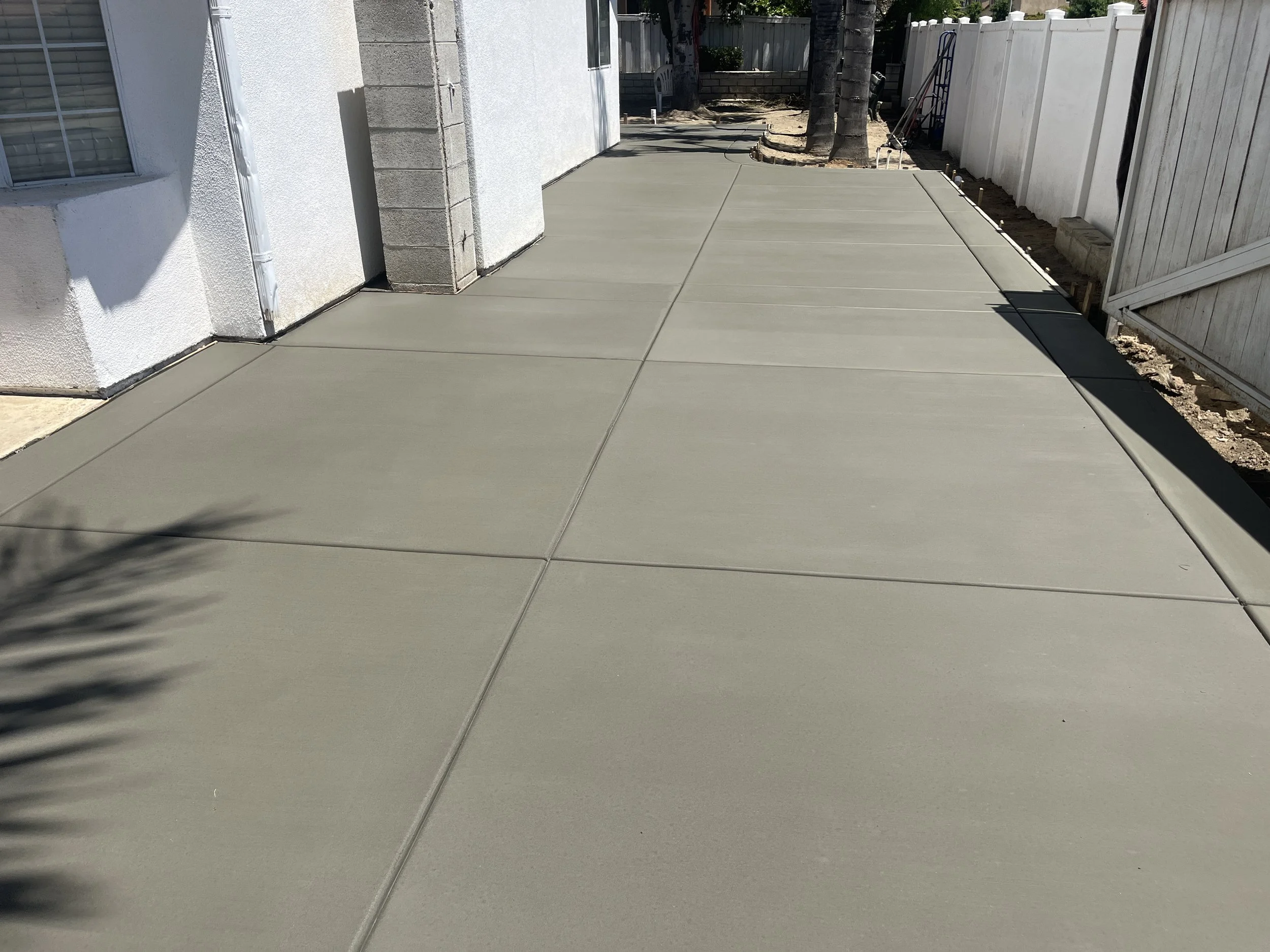 Freshly poured concrete sidewalk in a backyard with a white fence on the right, a house on the left, and some trees at the far end.