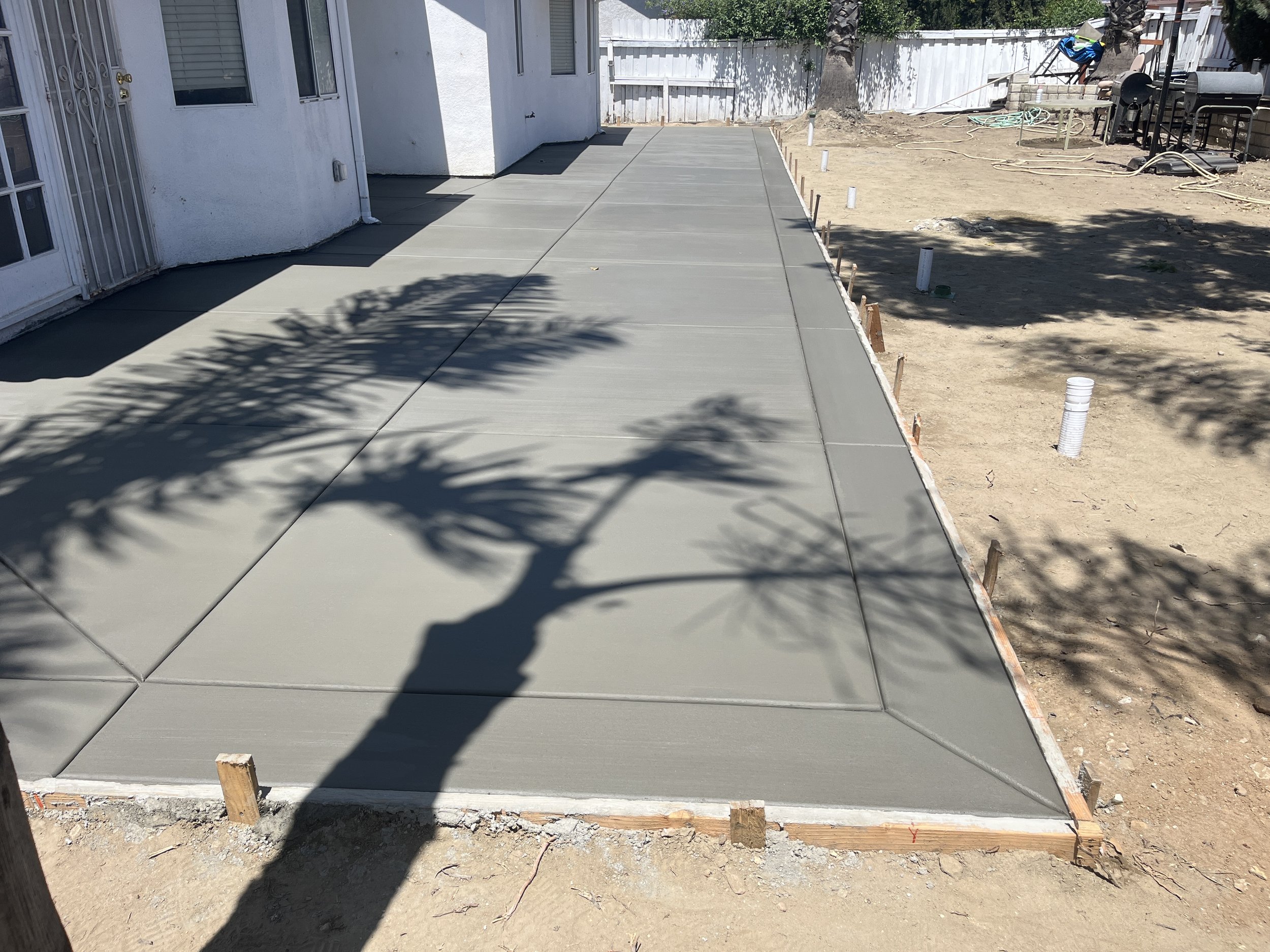 Freshly poured concrete sidewalk with wooden borders, adjacent to a white house with a glass door and window, shadows of trees cast across the surface, and surrounding area of bare dirt and construction materials.