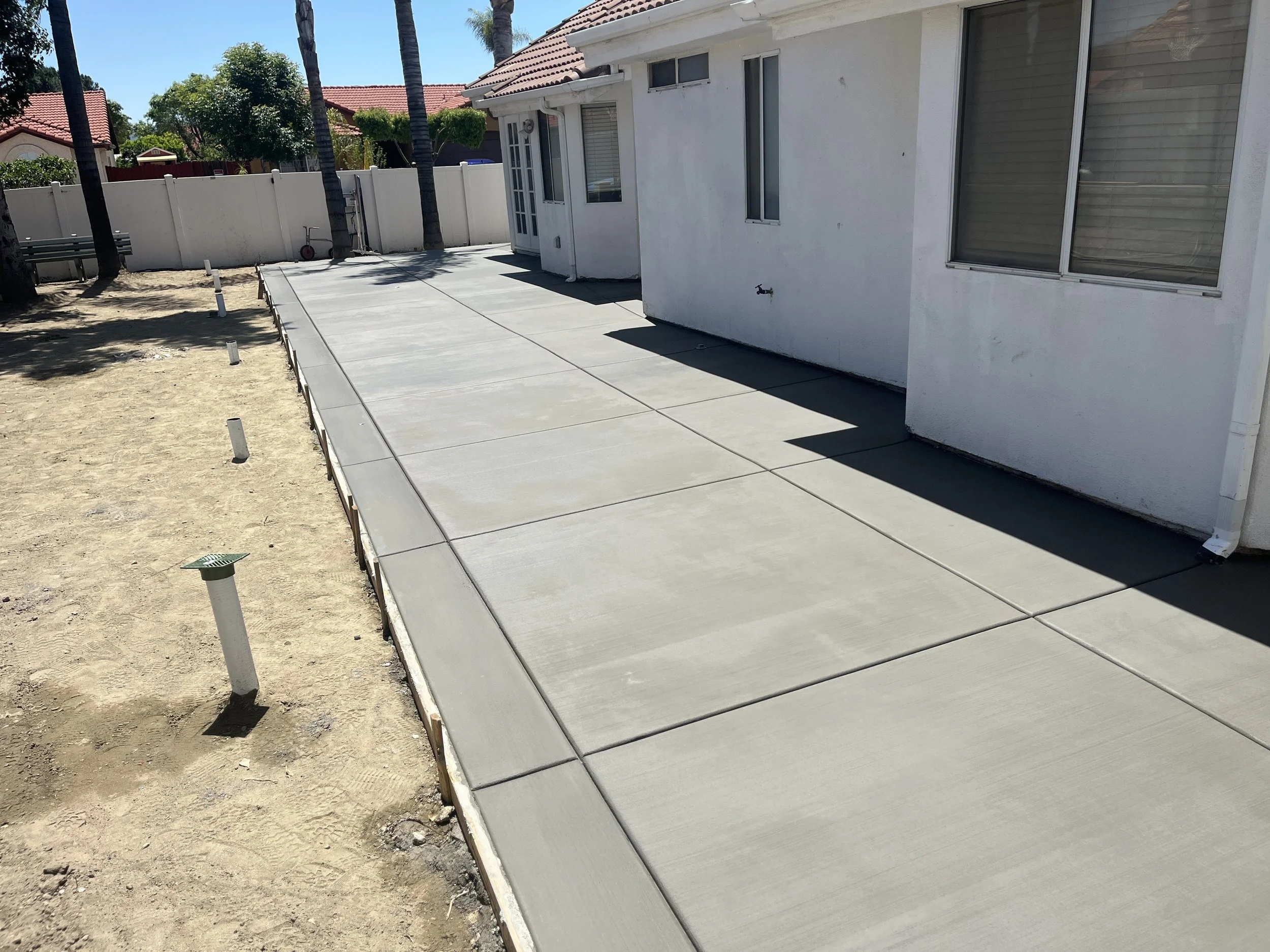 Newly poured concrete sidewalk next to a white house on a sunny day, with some soil and ground preparation visible on the side.