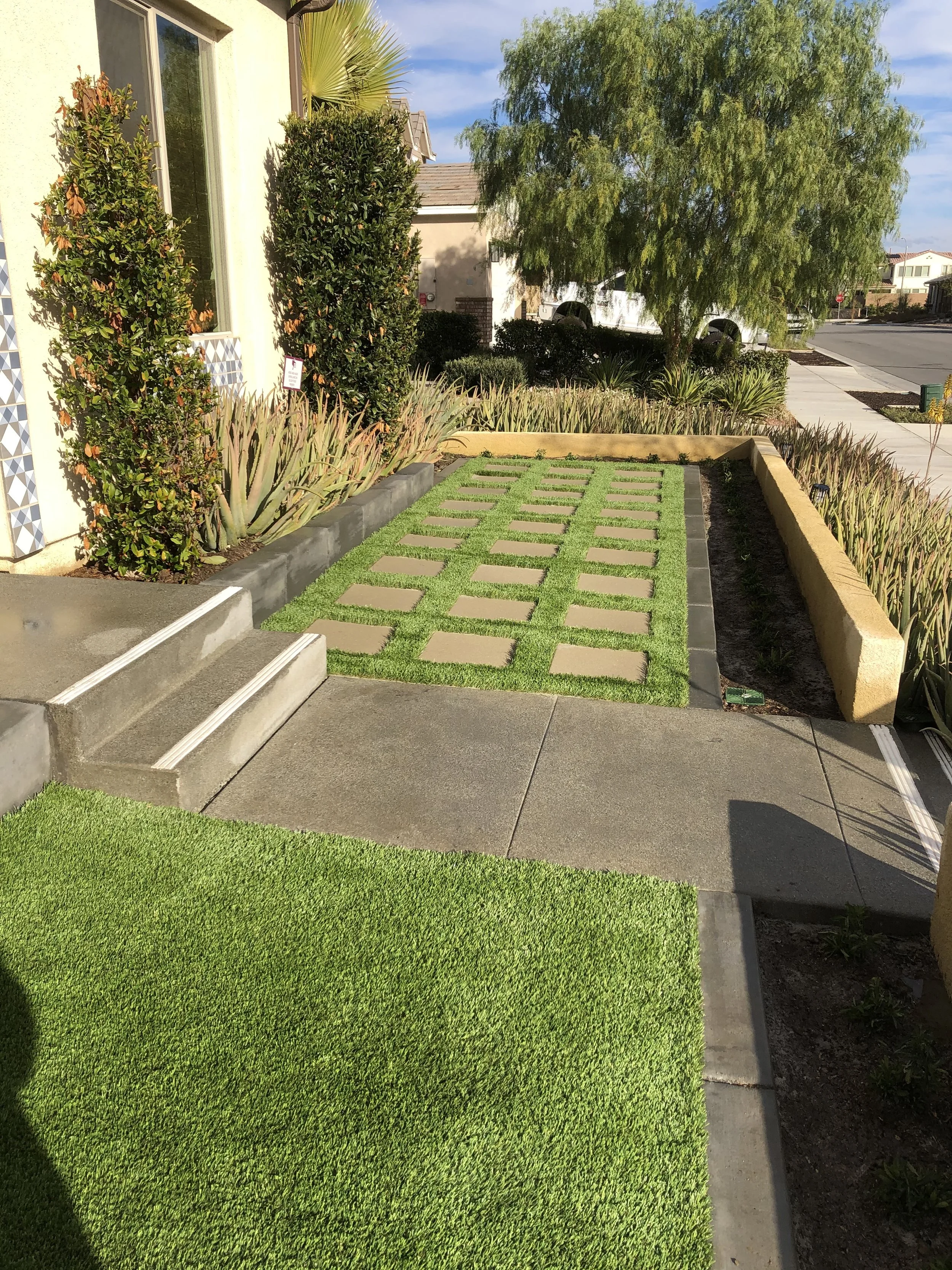 A neat residential front yard with a modern concrete walkway, a patch of artificial grass, decorative paving stones, and landscaped garden beds with green plants and shrubs, bordered by a low yellow wall on the right side.
