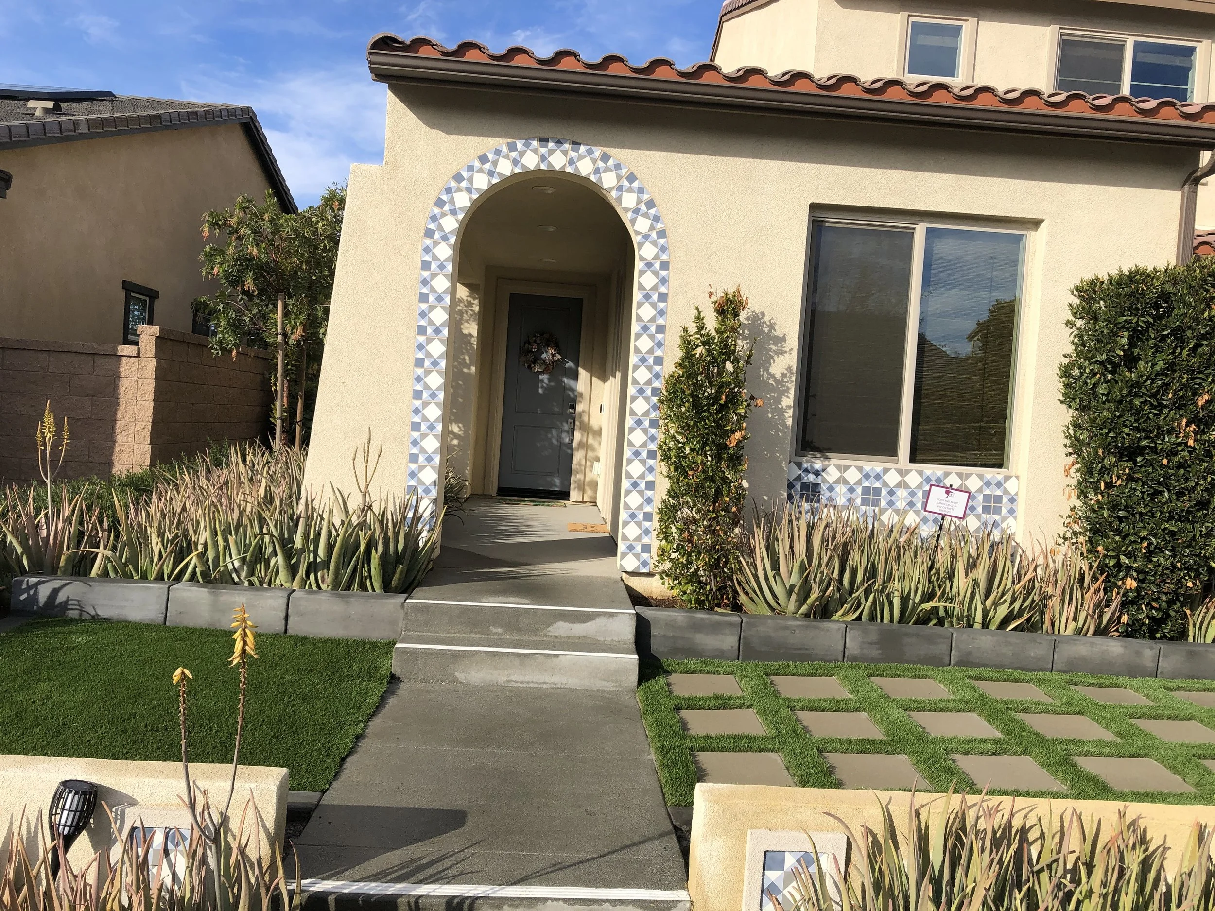 Front view of a modern house with a beige facade, a front door decorated with a wreath, and a curved archway with blue and white tiles. The front yard has green grass, succulents, and decorative concrete pavers.