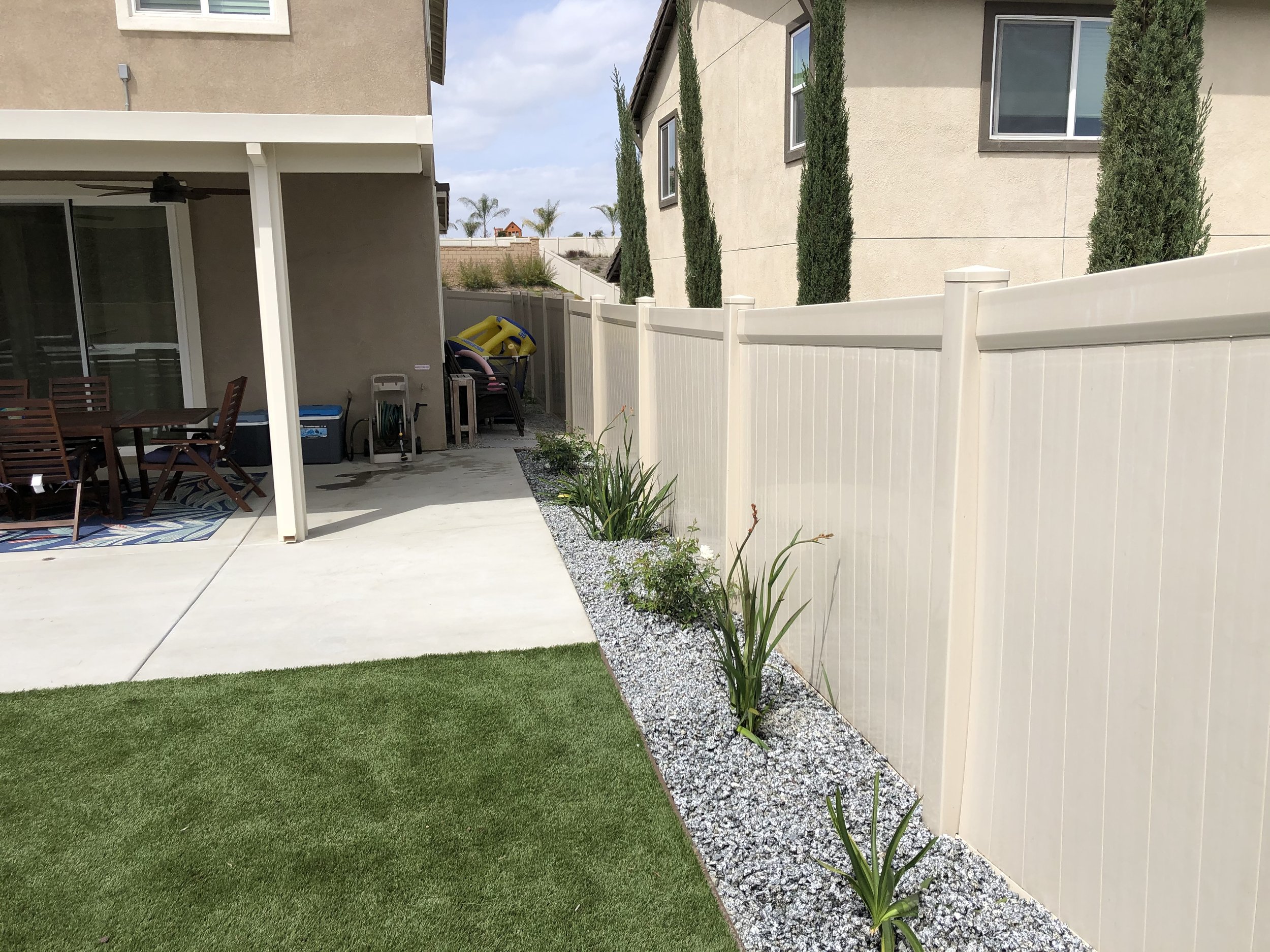 Backyard with a grassy lawn, concrete patio, beige privacy fence, and a row of tall, narrow cypress trees.  Decorative concrete and aloe landscape design – Menifee, CA