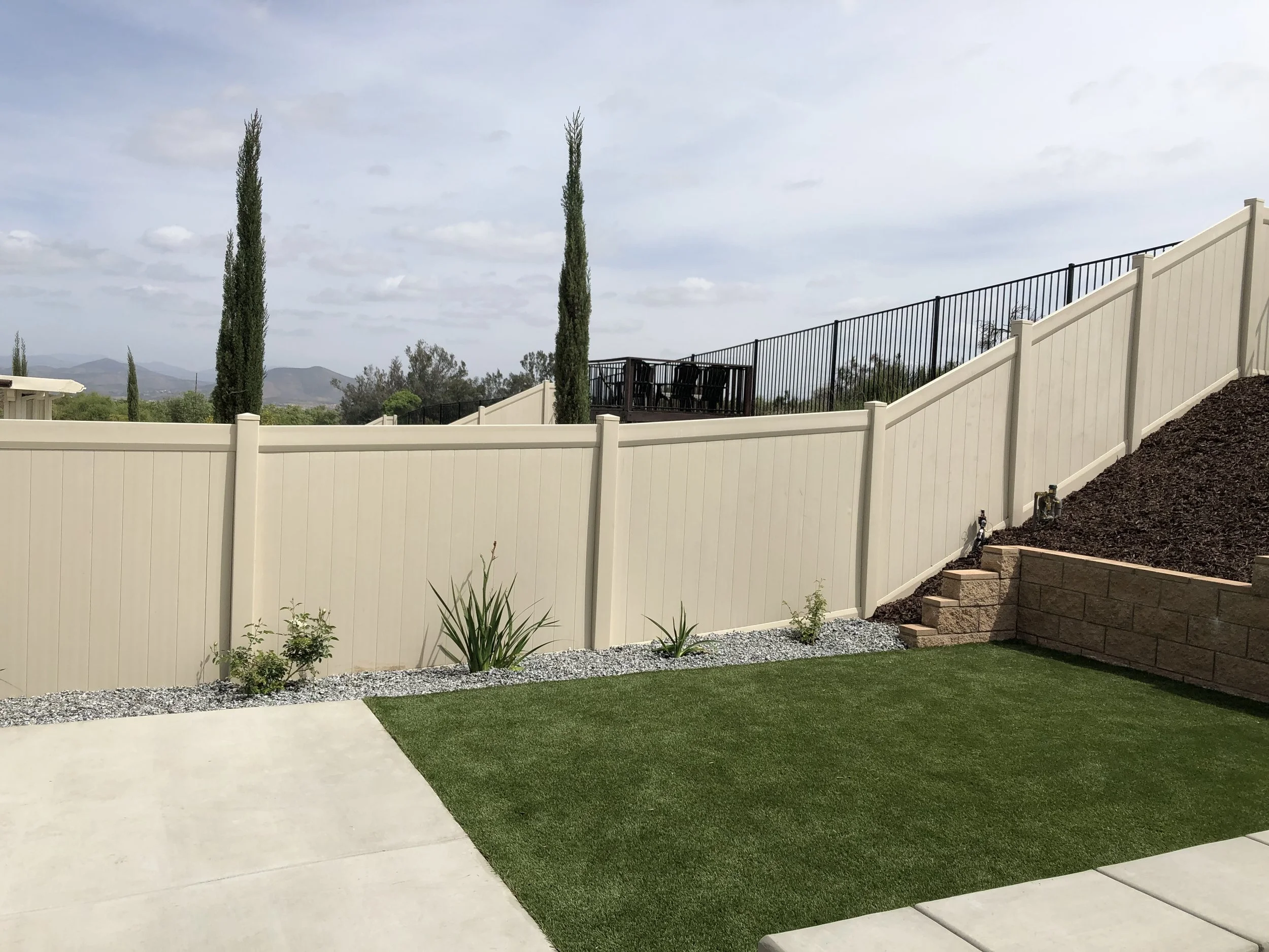 Backyard with neatly mowed grass, a beige vinyl fence, and small plants along the fence. The yard is on a sloped hill with stairs and mulch on the right. In the background, there are two tall, thin cypress trees and a view of distant mountains under 