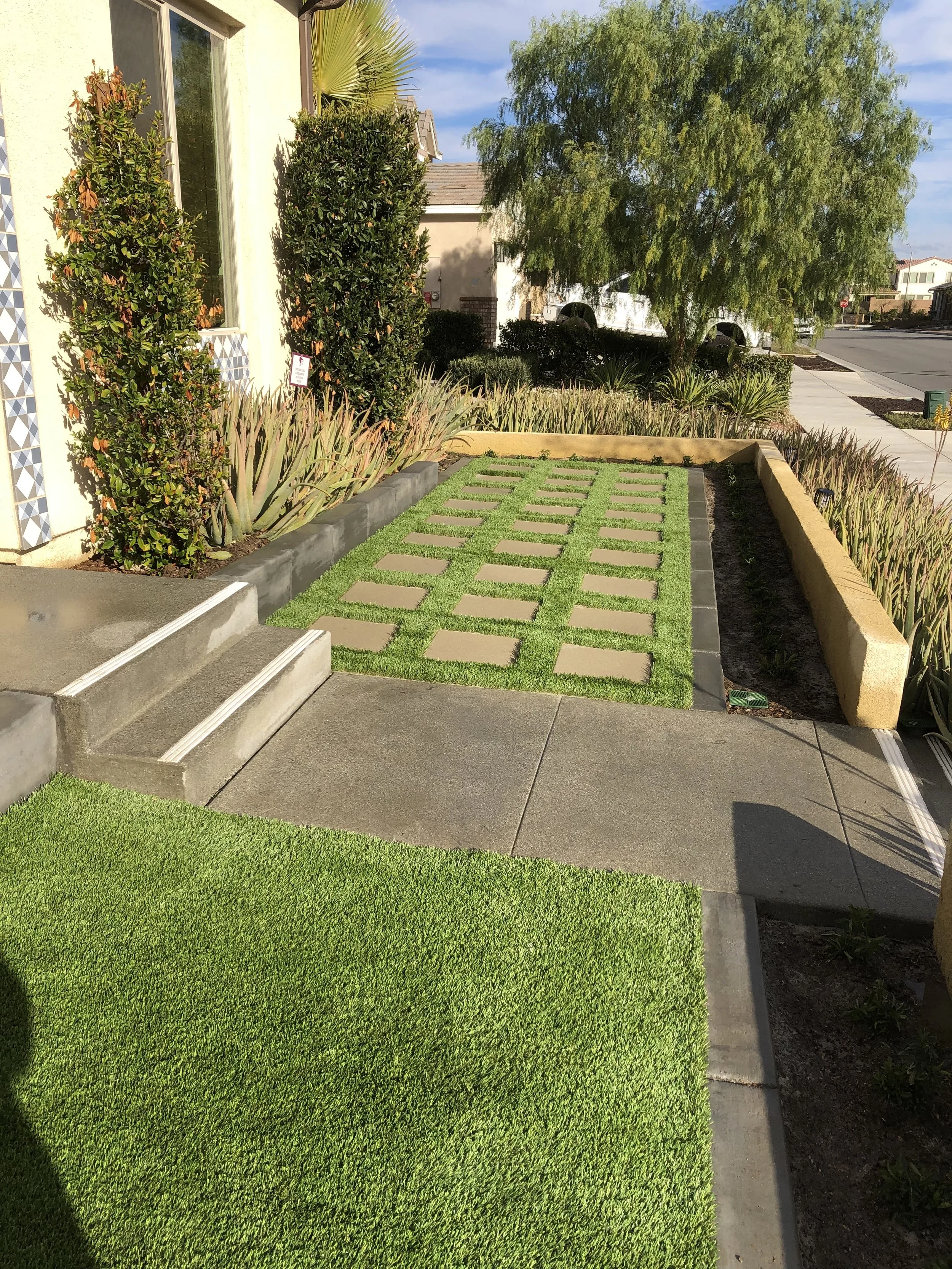 A front yard with a small grassy lawn, concrete steps leading to a door, a pathway with square stepping stones, landscaped bushes, and a tree near the sidewalk.