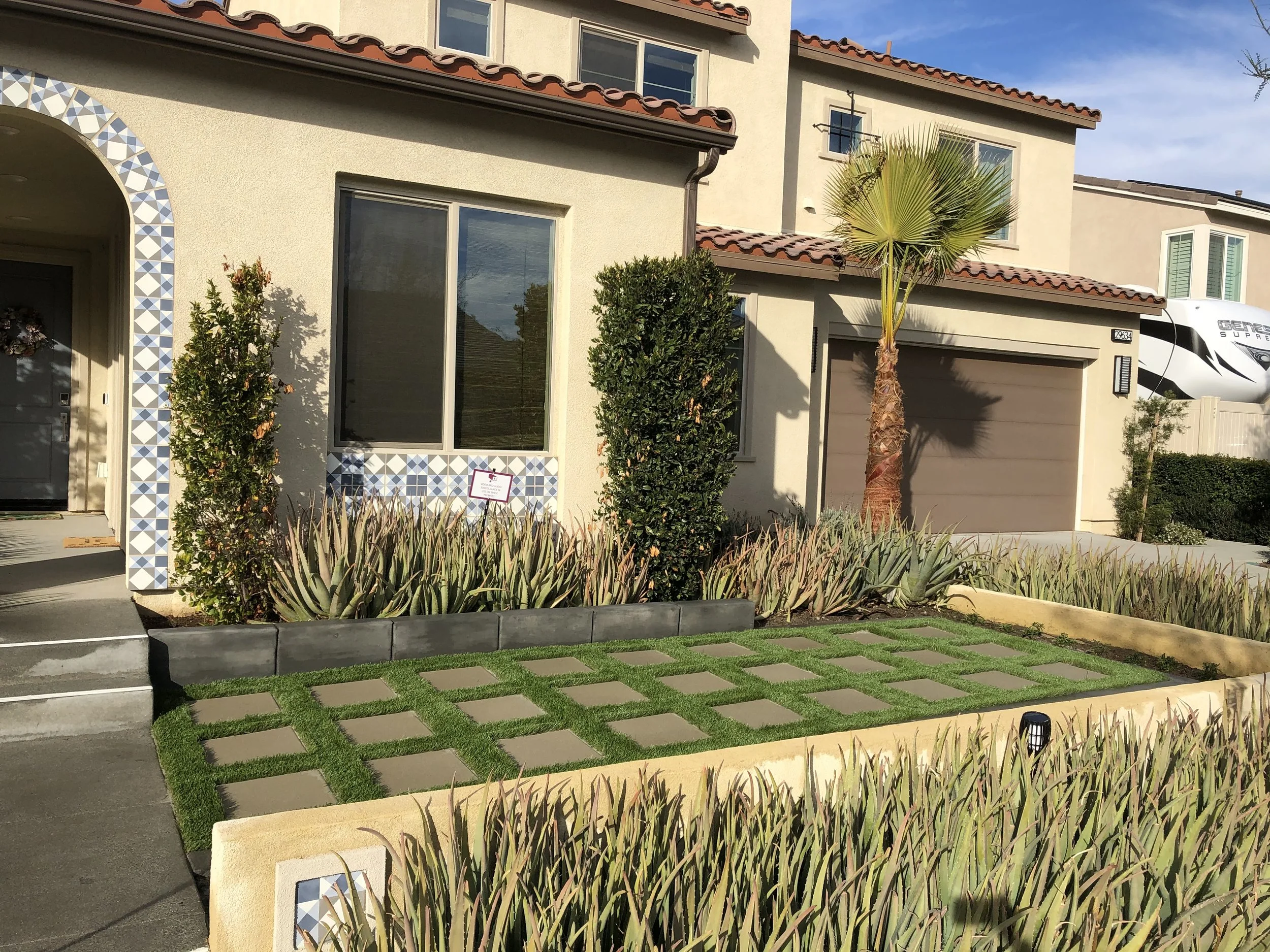 Front yard of a house with a small lawn, decorative plants, a palm tree, and a window with geometric tile border.