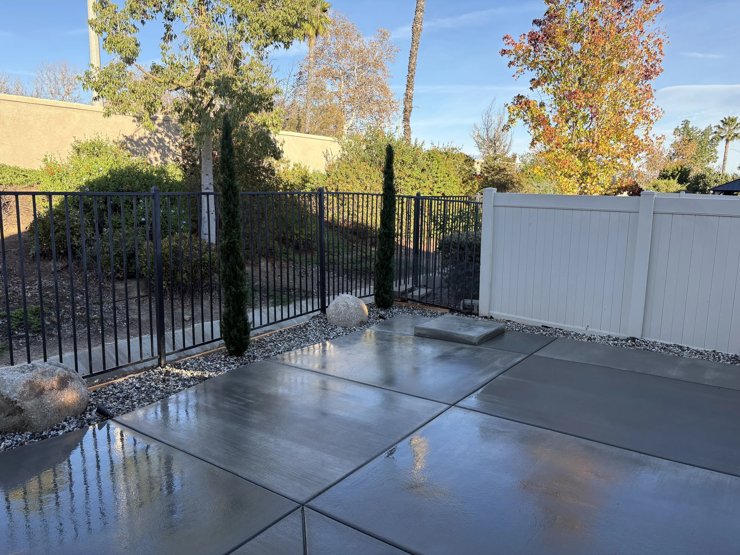 View of a backyard patio with concrete slabs, white fence to the right, black metal fence to the left, and trees with fall foliage in the background.