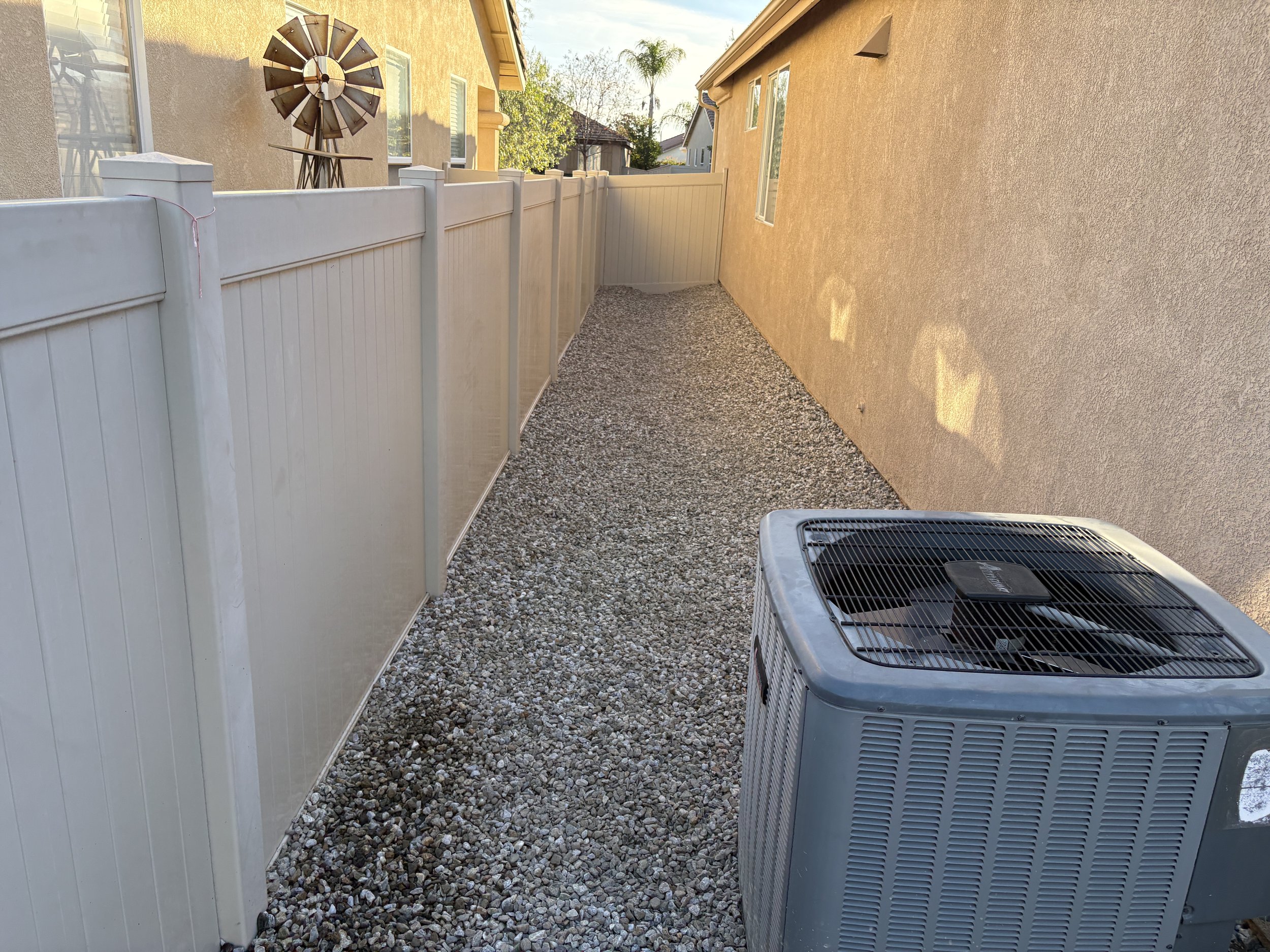 A backyard alleyway with a beige wall on the right and a white vinyl fence on the left. The ground is covered with small gravel. An air conditioning unit is in the foreground on the right. In the background, there is a windmill decoration mounted on 