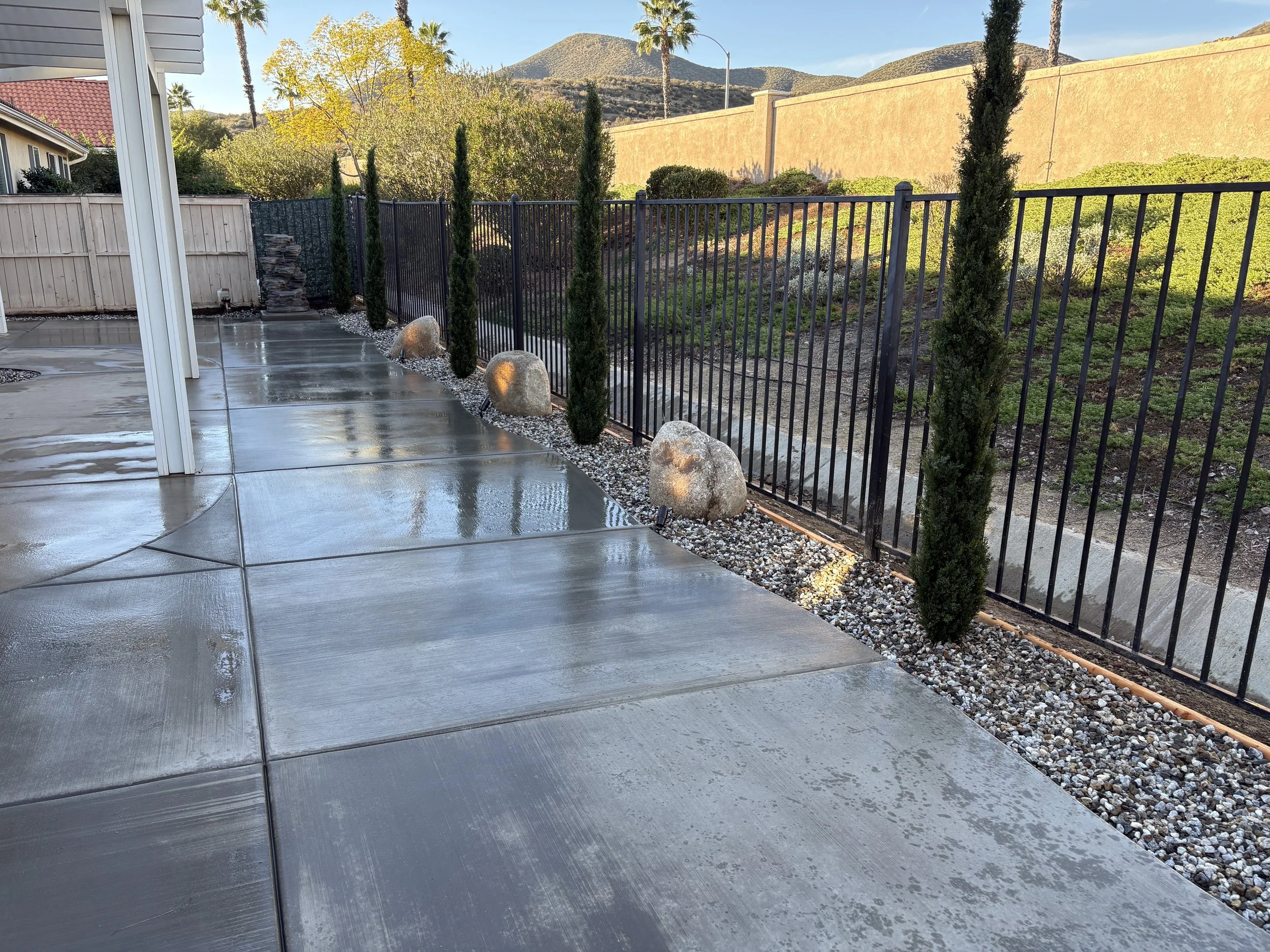 A wet concrete patio with a black metal fence, tall slender cypress trees, decorative rocks, and a view of hills and trees in the background under a clear blue sky.