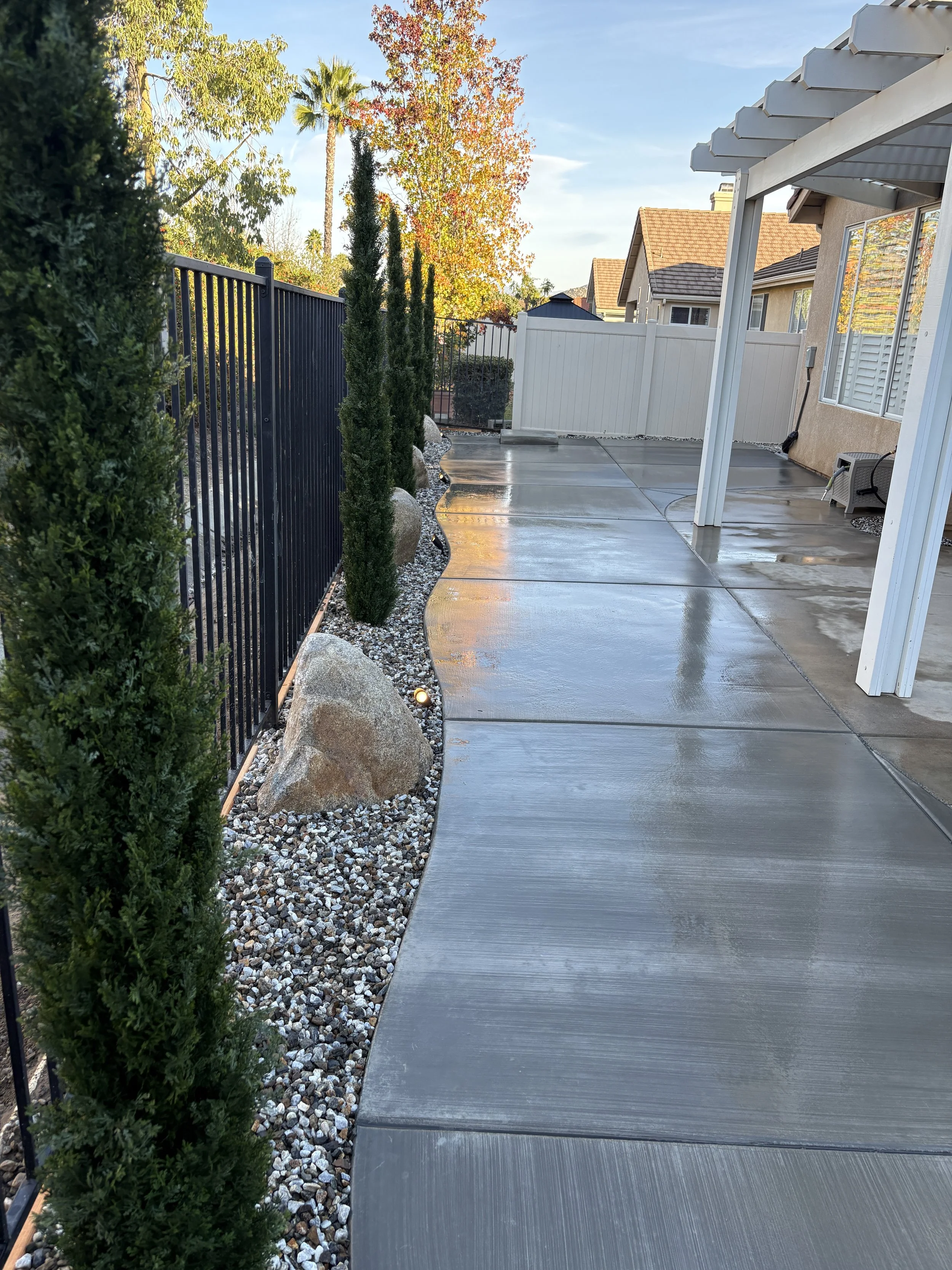 Clean concrete patio with a white fence, tall trees, and a patio cover, Checkerboard concrete pattern with custom wavy edges installed in Murrieta, CA.