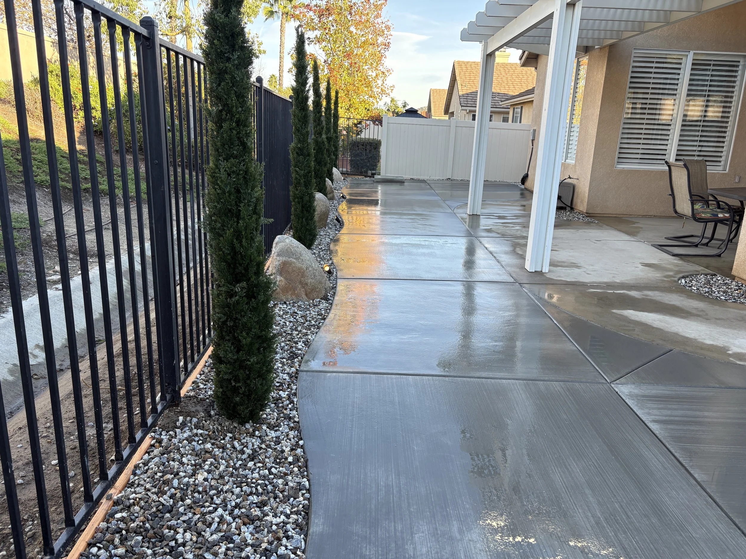 Wet concrete patio with a black metal fence, decorative rocks, and small trees along the edge of a backyard patio. Custom Murrieta landscape project featuring decorative concrete, boulders, and water feature.