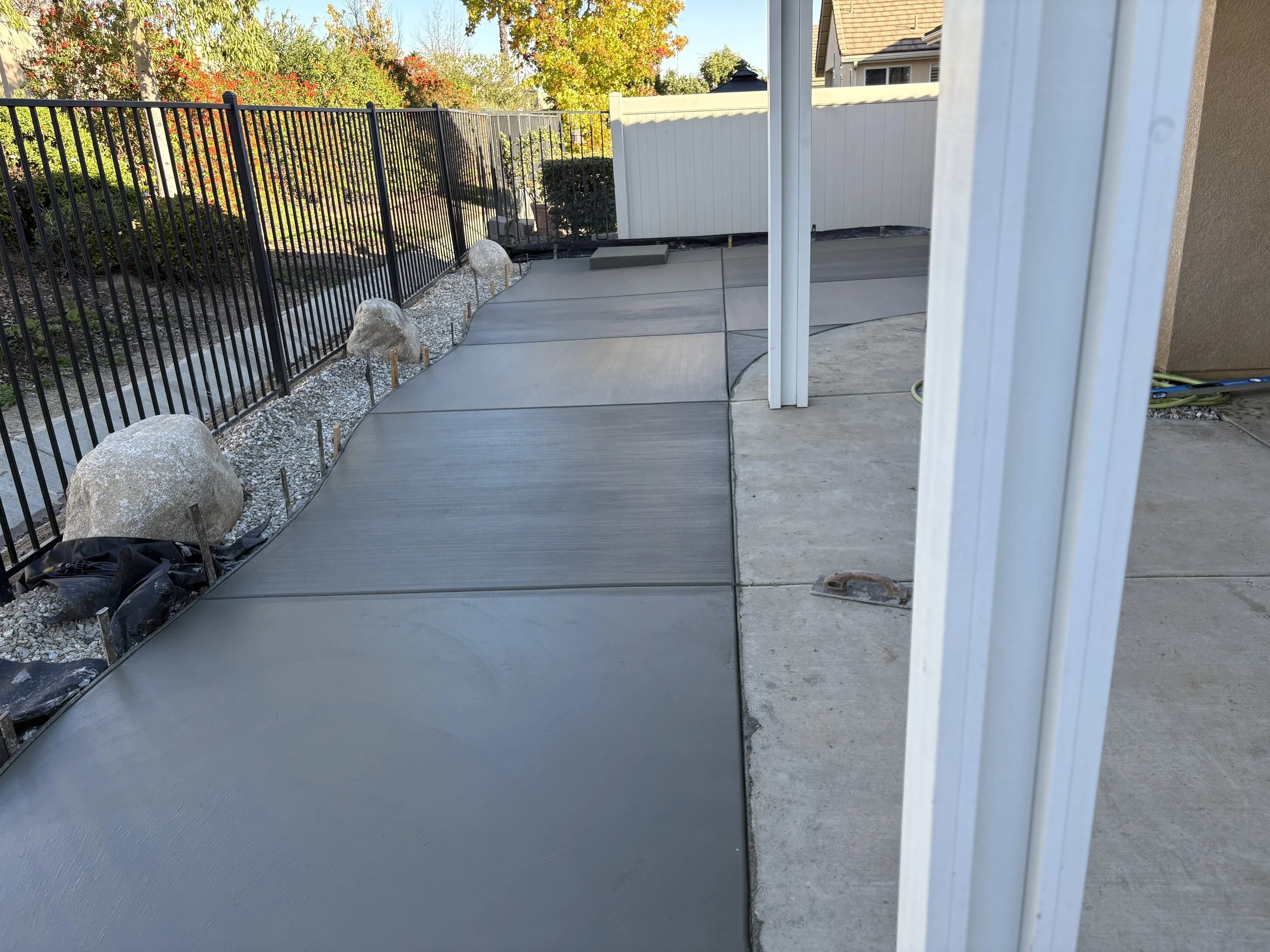 Newly poured concrete sidewalk with curves, bordered by rocks and a black metal fence on the left, and a concrete patio on the right. A white fence and trees with autumn foliage are in the background.