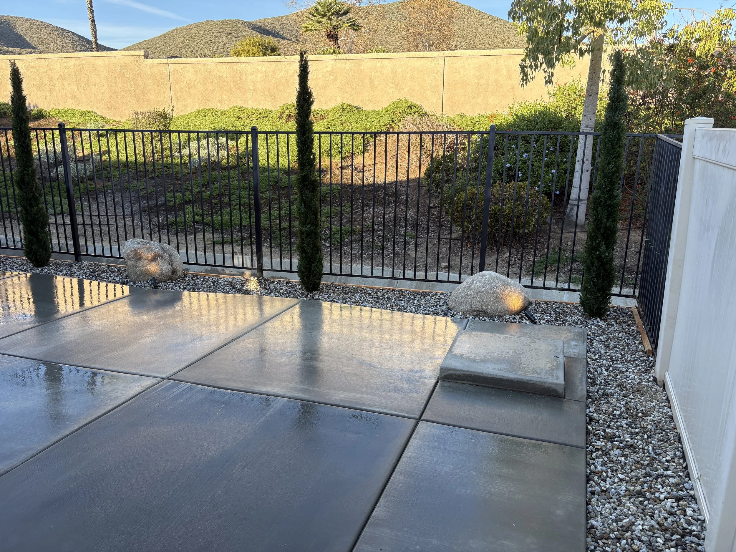 A backyard patio with wet concrete tiles, black metal fence, small cypress trees, large decorative rocks, and a beige privacy wall with mountains and trees in the background.