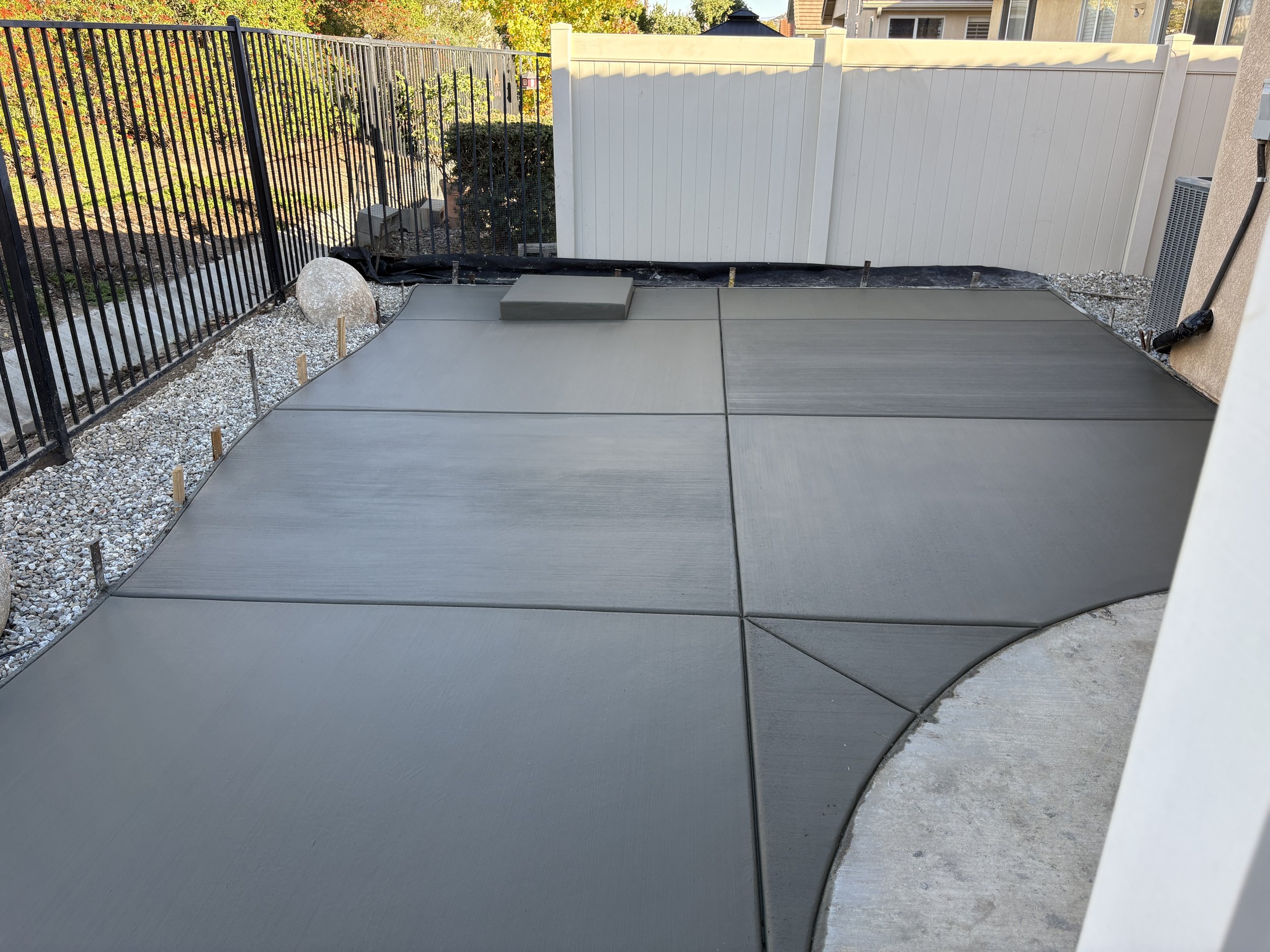 Freshly poured concrete patio with sealing joints, bordered by a curved edge, with a black metal fence on the left and a white fence in the background, and neighboring house wall on the right.