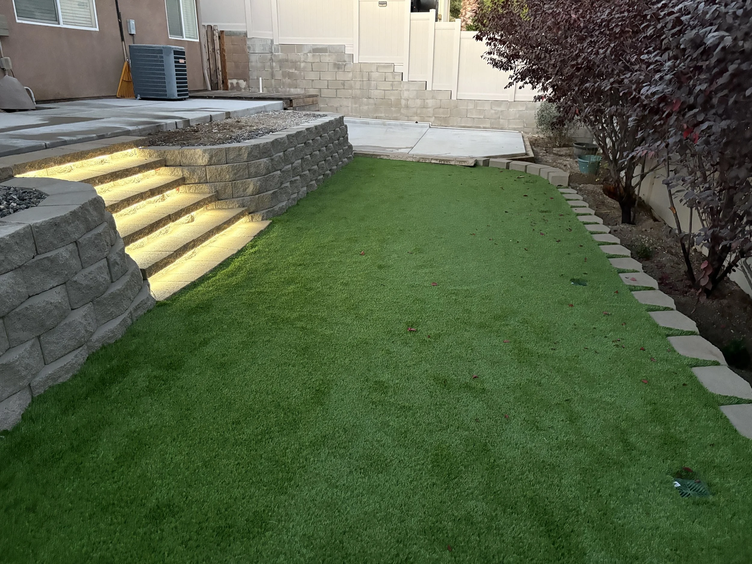 Backyard with green artificial turf, stone steps with built-in lights, a retaining wall with gravel, and a planted area with bushes along the right side. Illuminated stone steps and retaining wall – Canyon Lake, CA