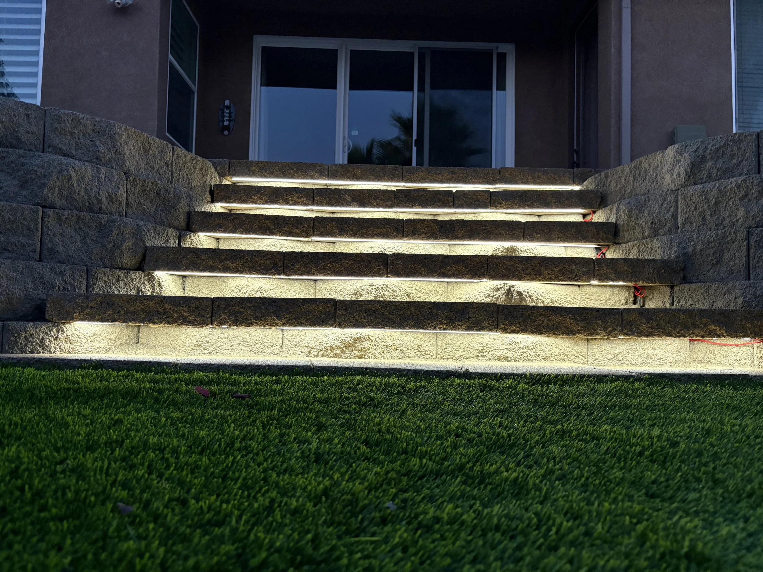 Outdoor staircase leading up to a house, illuminated with built-in lighting, with a grassy lawn in the foreground.