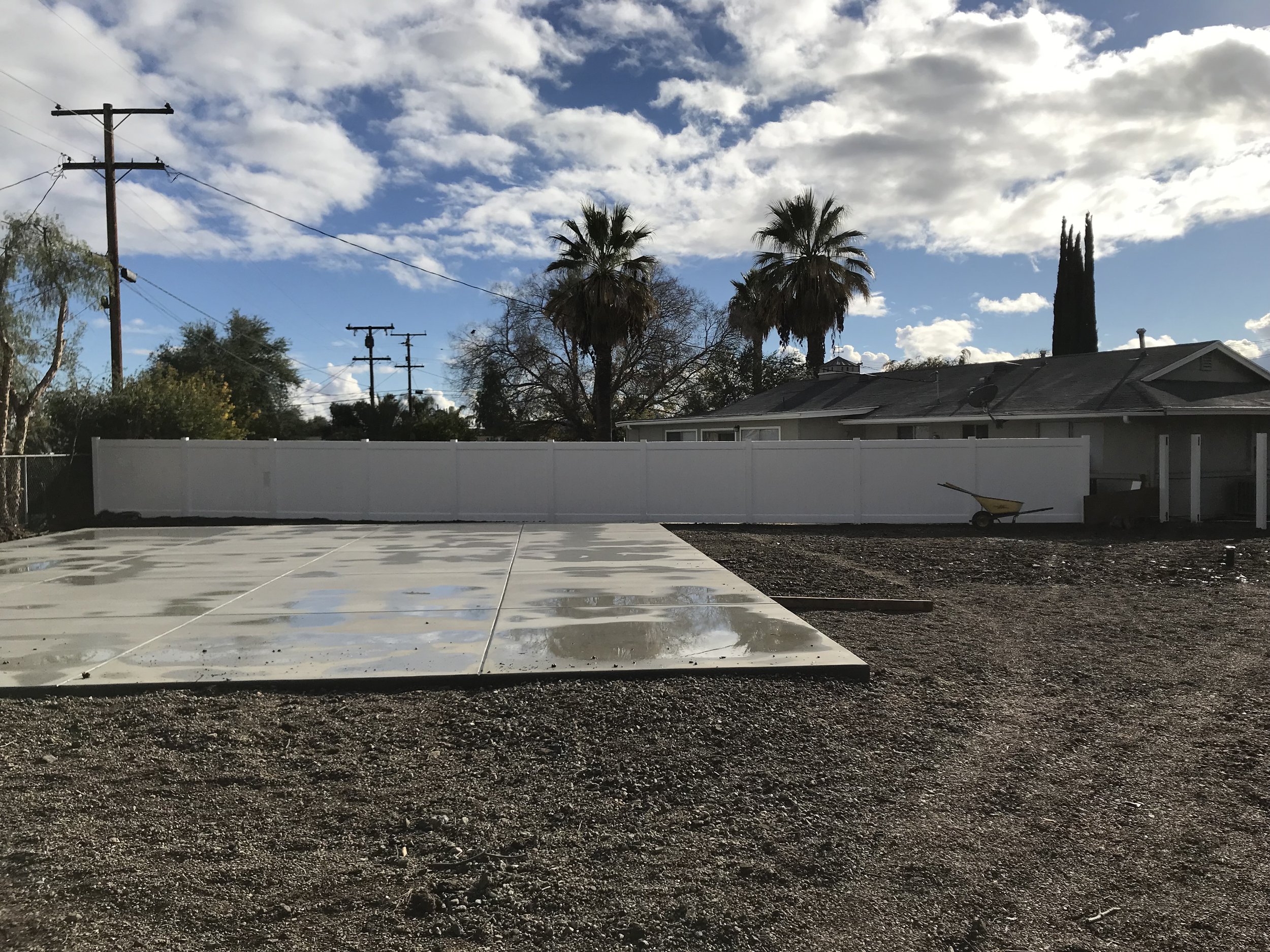A backyard with a concrete pad, a white privacy fence, two palm trees, and a cloudy sky.