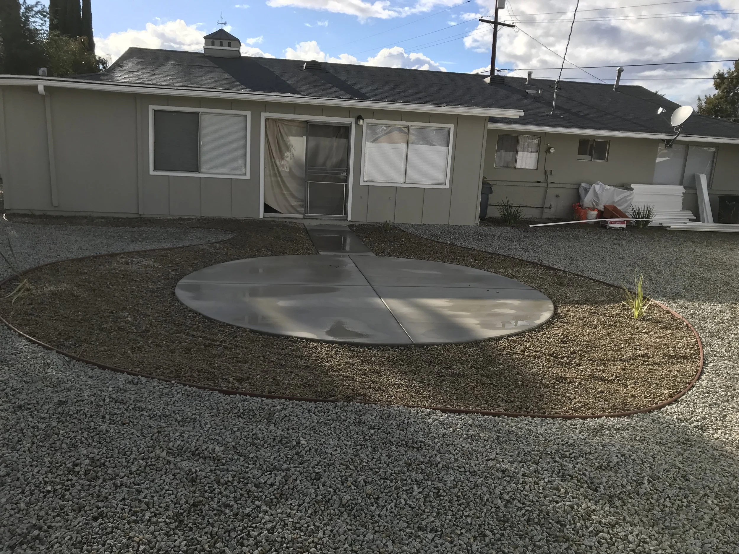 A backyard with a gravel ground, a circular metal cover, and a small garden bed with a plant, with a house in the background that has a screened door and windows.
