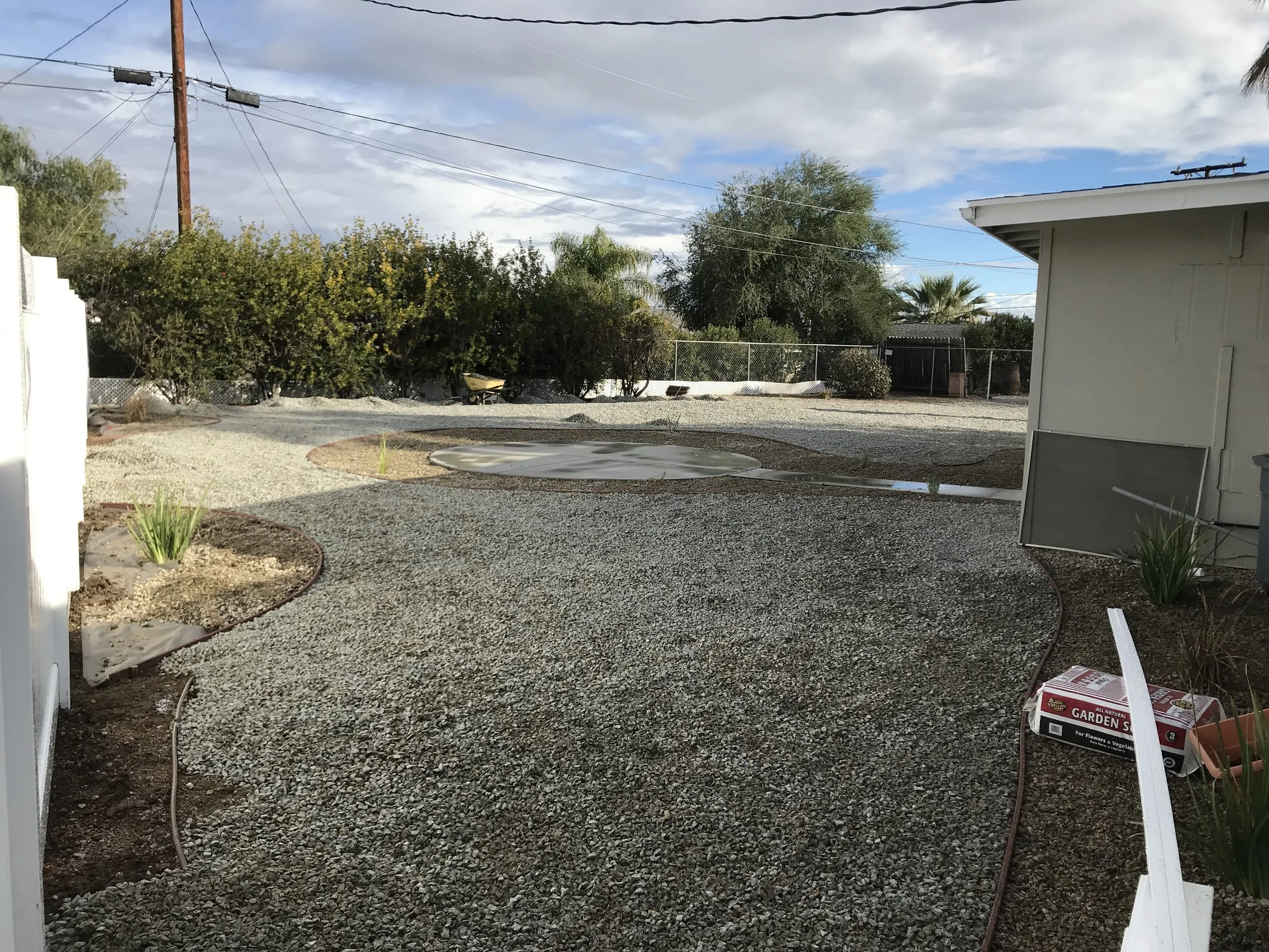 A backyard with a gravel pathway, small garden beds with plants, a white fence on the left, bushes and trees in the background, and a grey shed on the right. The sky is partly cloudy with patches of blue.