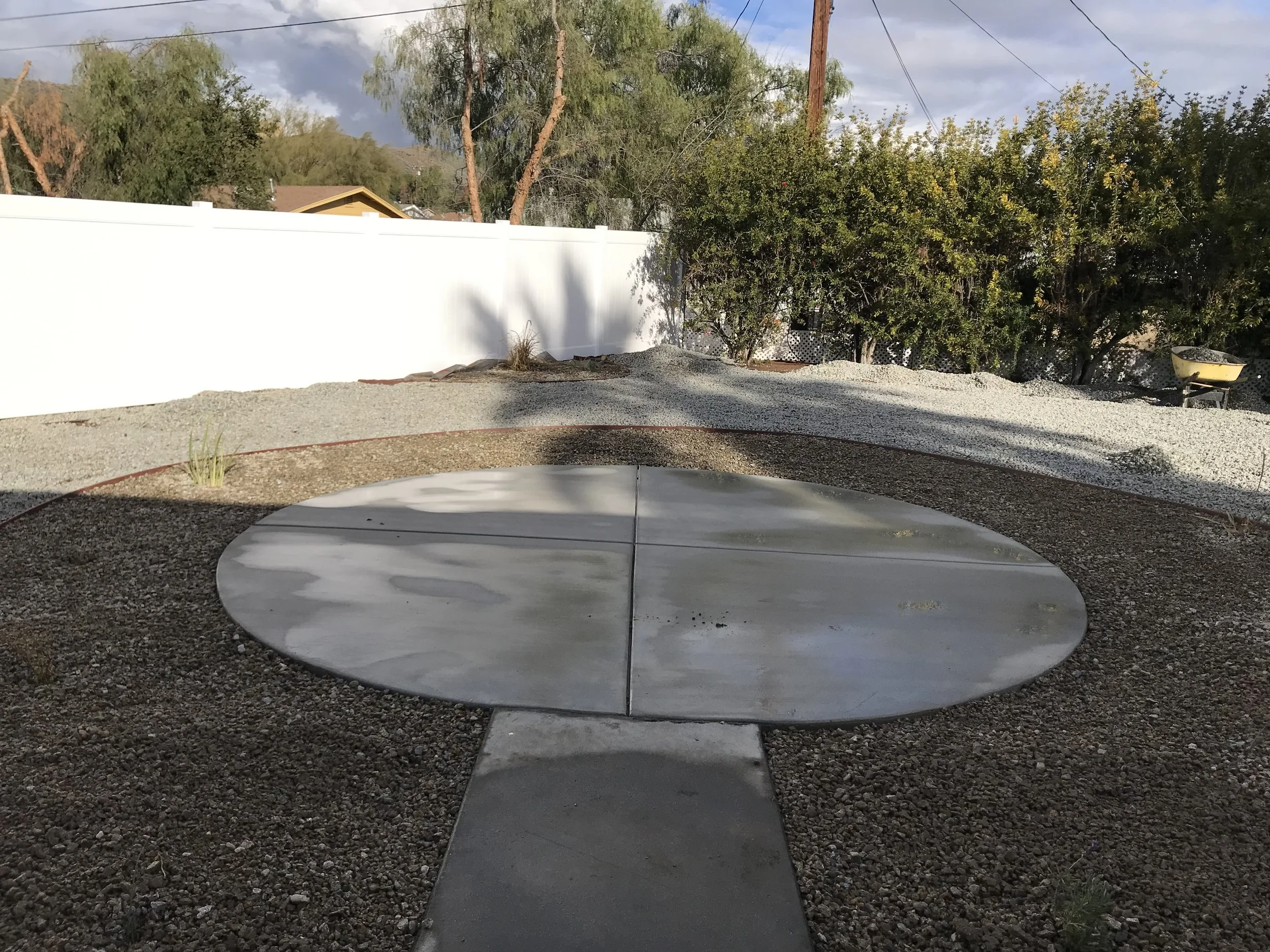 A backyard with a circular concrete pad, a white fence, green bushes, and trees under a partly cloudy sky.