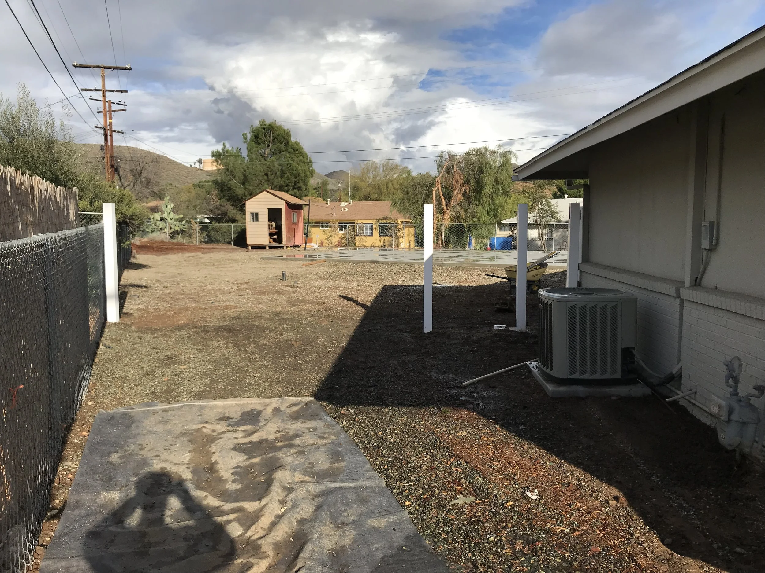 Backyard with a gravel and dirt area, a small wooden playhouse in the distance, metal chain-link fence, a house with an air conditioning unit, and a cloudy sky.