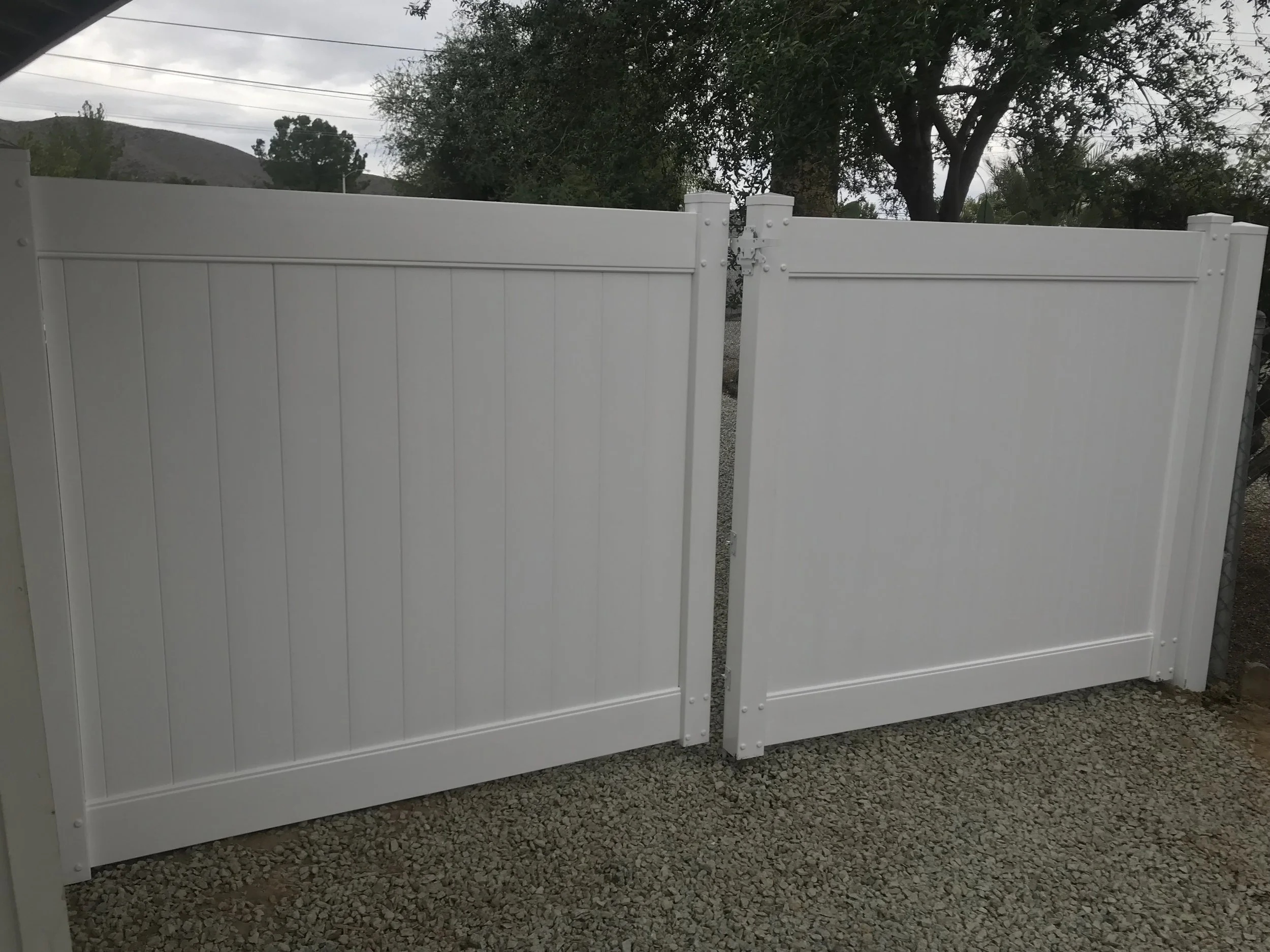 White double gate fence with vertical panel design, situated over gravel ground, with trees and power lines in the background.