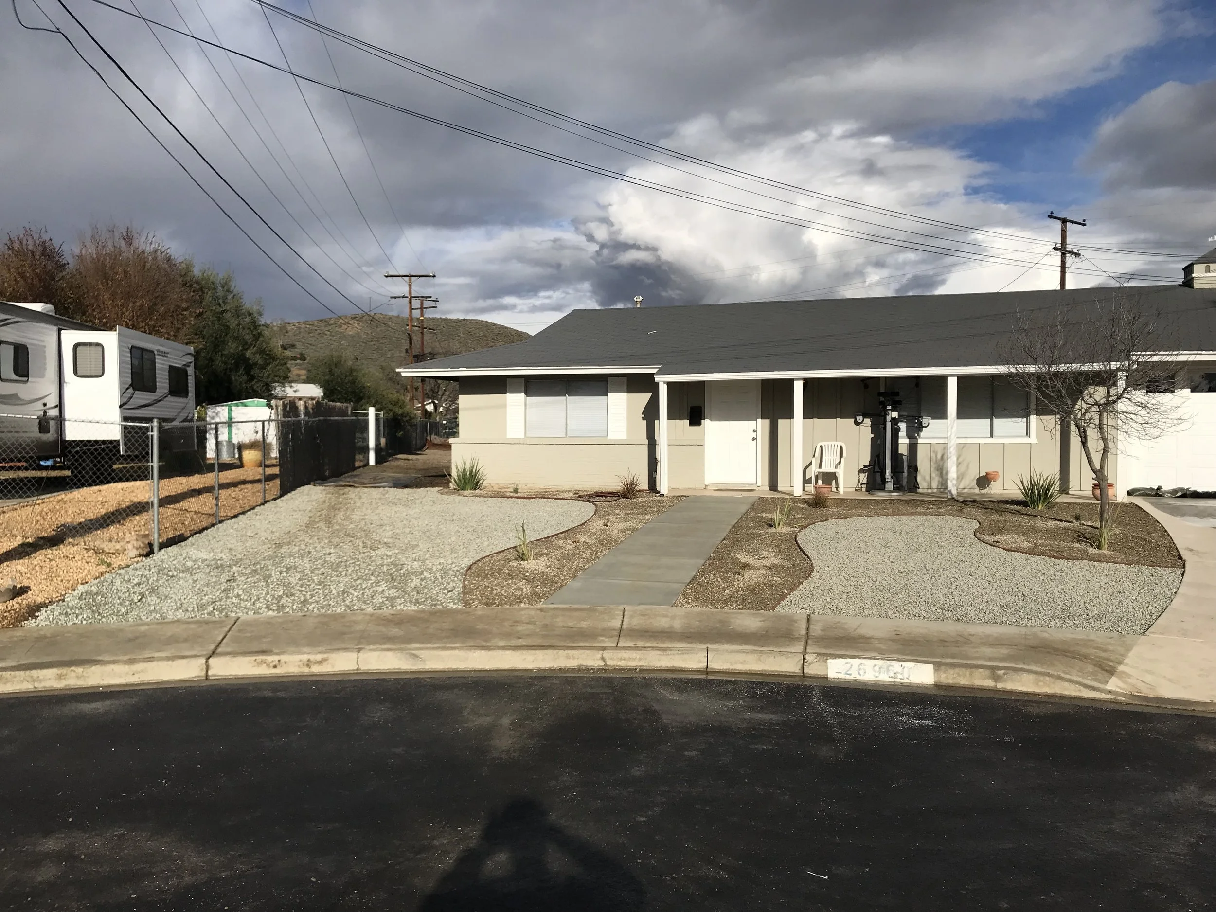 Front view of a house with a garden that has gravel patches and a walkway, a white plastic chair, exercise equipment outside, and a tree with no leaves, under a partly cloudy sky.