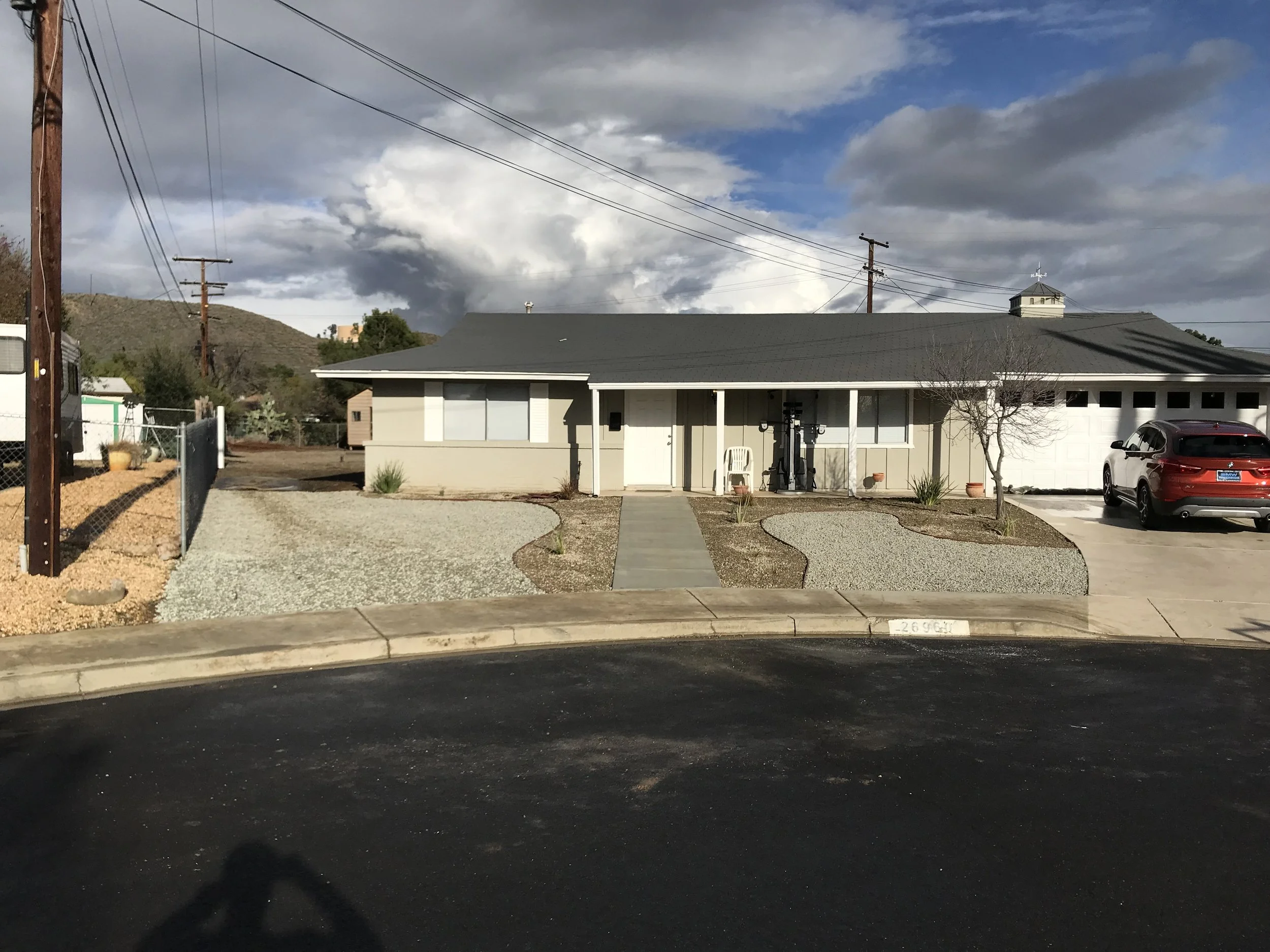 Single-story house with beige exterior, black roof, front porch with white chair, small landscaping with grass and gravel, leafless tree, parked white SUV, and cloudy sky.