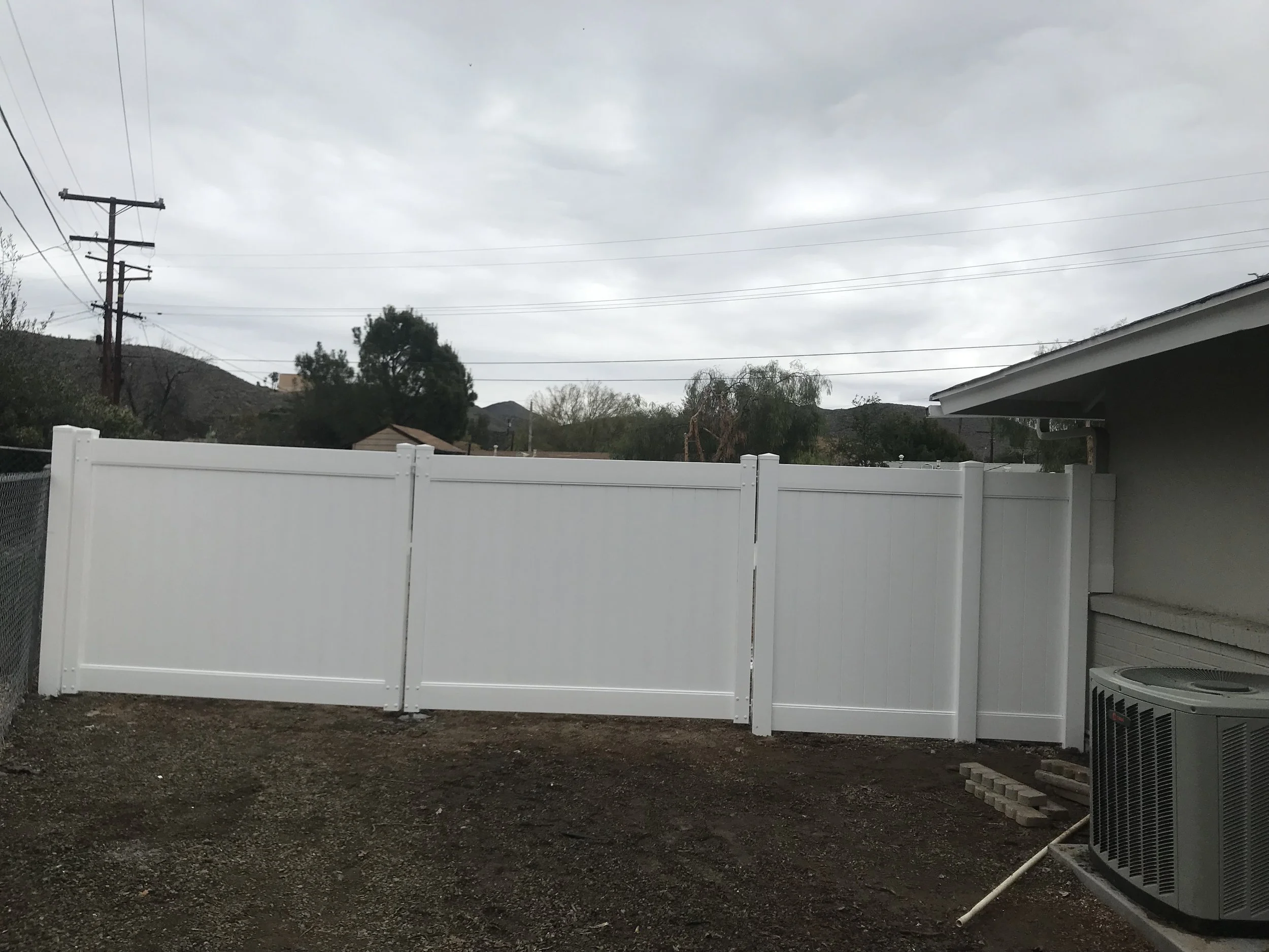 A backyard with a white vinyl privacy fence, an air conditioning unit, and a partly cloudy overcast sky. There are mountains and power lines in the background.