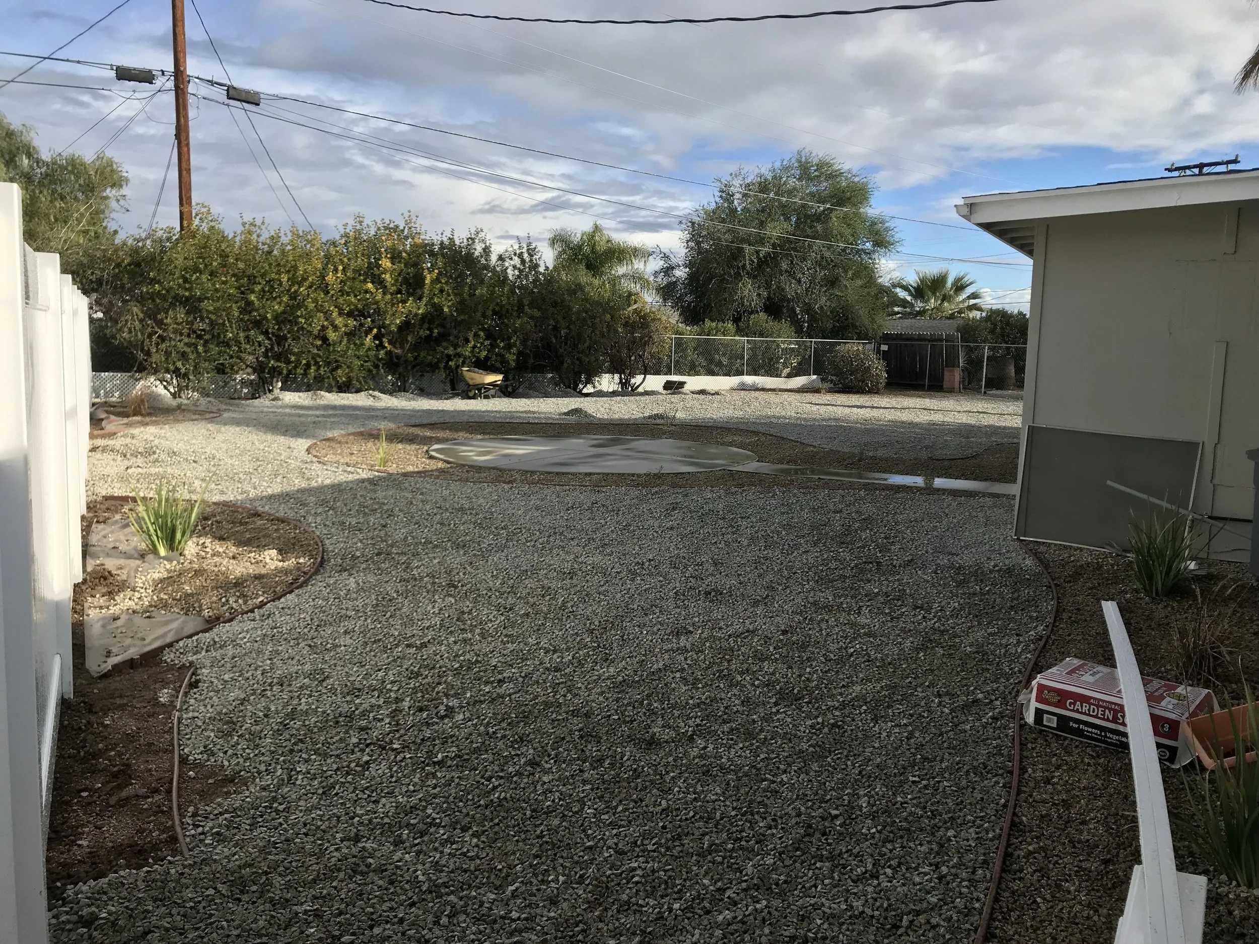 Backyard with gravel ground, plant beds, a small pond, and trees in the background. There is a house on the right and gardening supplies on the ground.
