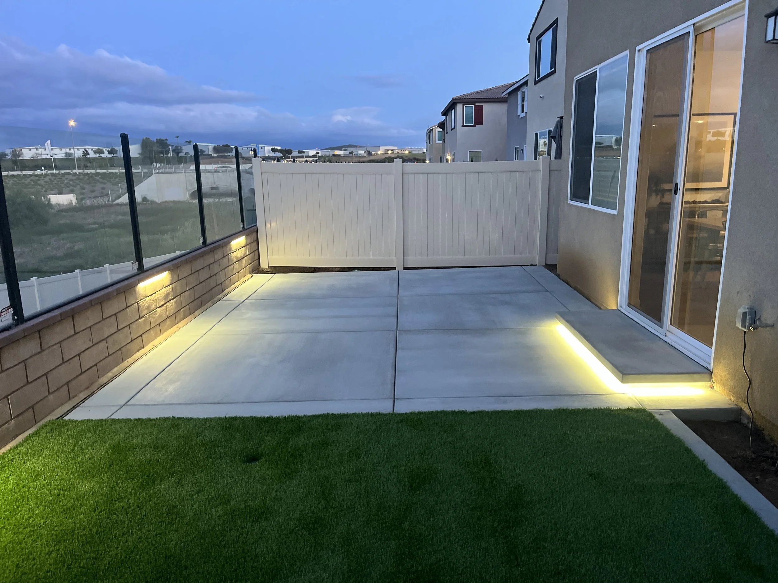 Backyard patio with concrete paving, artificial grass, a fence, and sliding glass door with outdoor lighting at dusk.