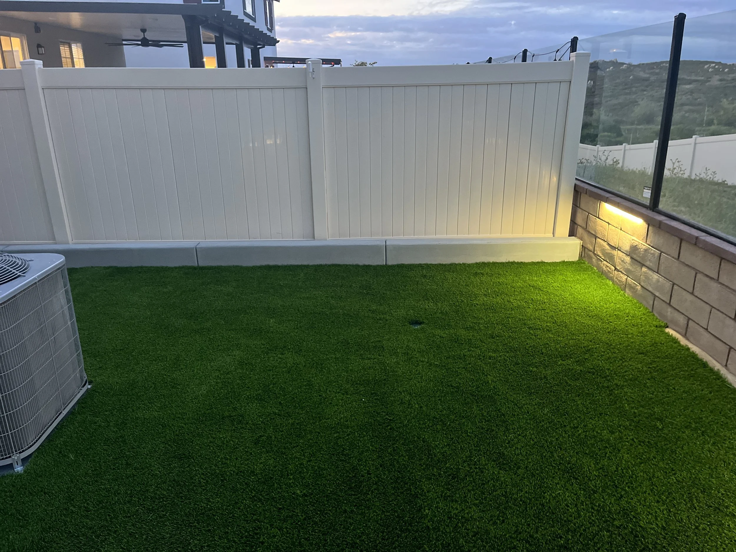 Small outdoor area with artificial grass, a white privacy fence, brick wall with a light, an air conditioning unit, and a glass barrier with a scenic view of hills in the background.
